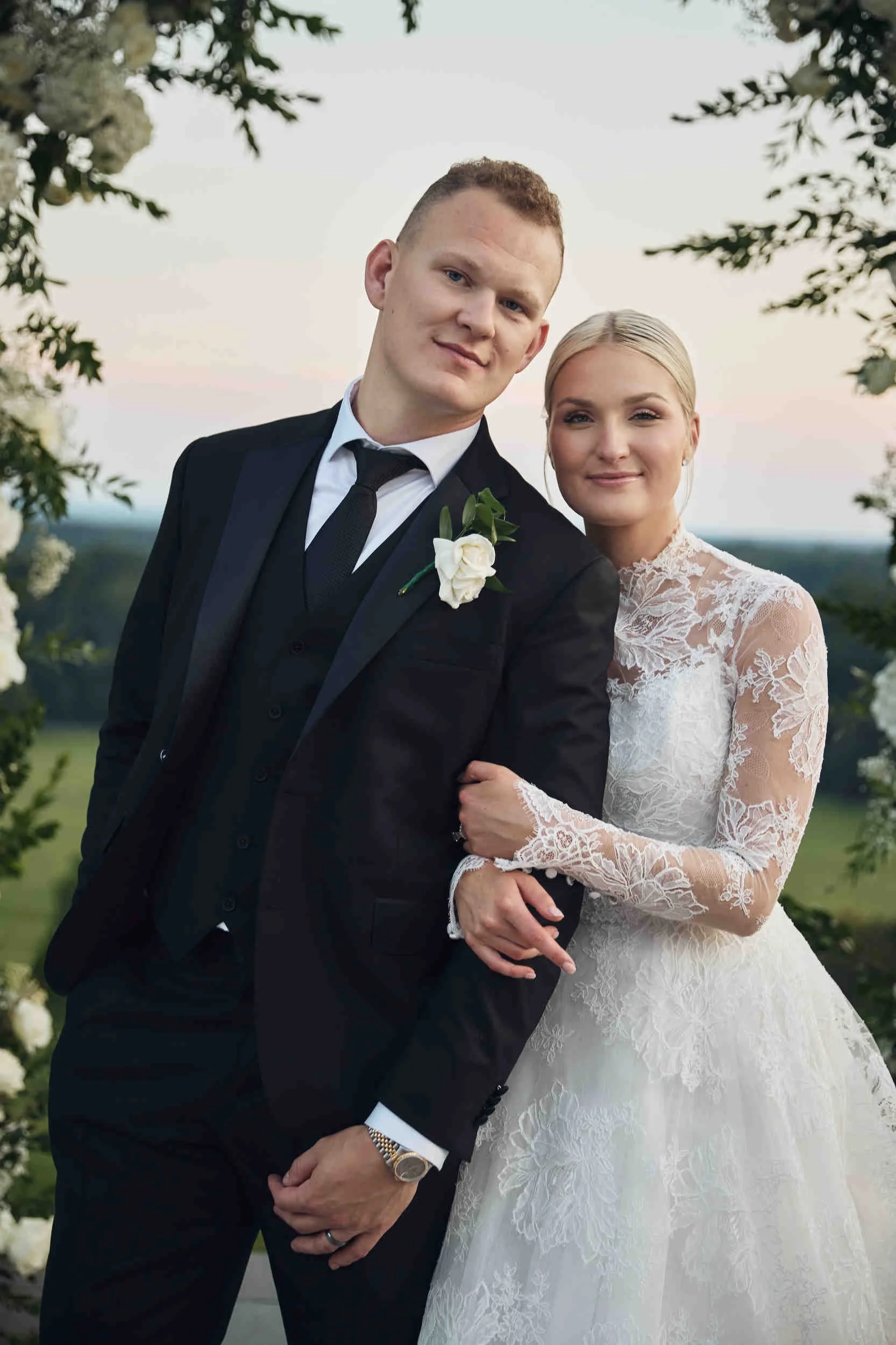 A newlywed couple dressed in wedding attire, standing outdoors with trees and a scenic landscape in the background, embracing each other and smiling.