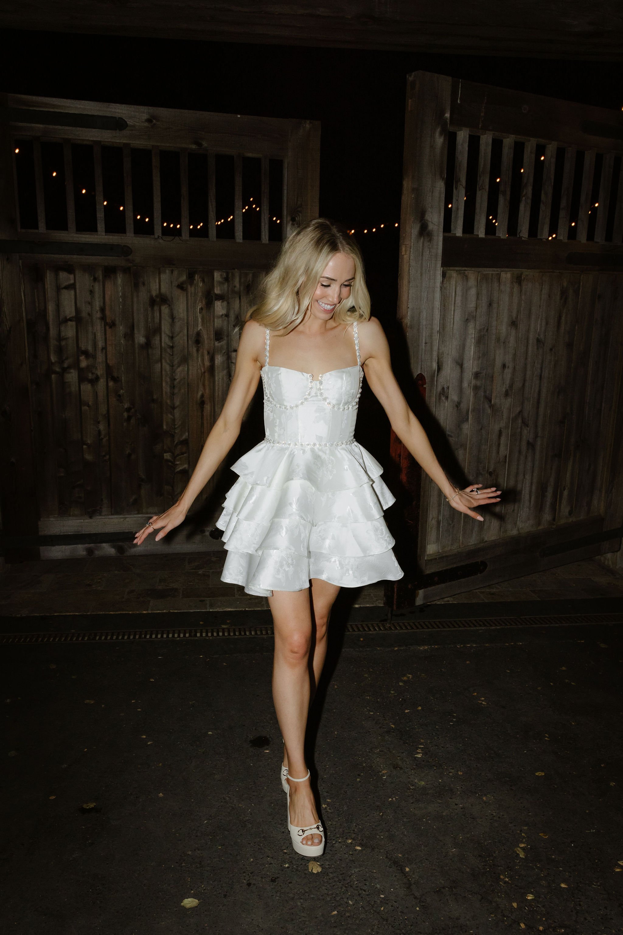 A woman in a white dress and high heels standing outdoors at night with a wooden fence and string lights in the background.