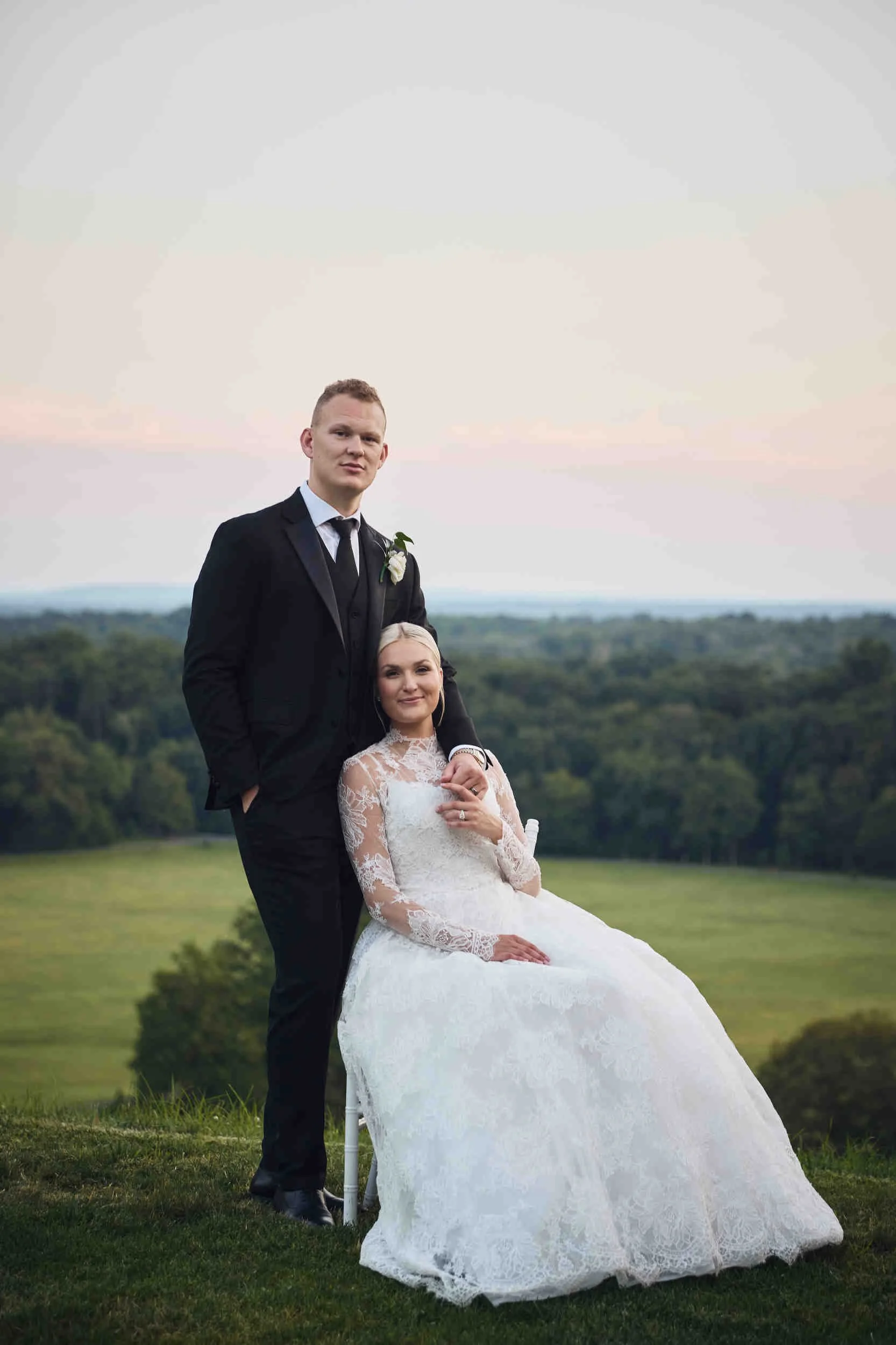 A bride and groom stand together outdoors, in formal wedding attire, against a scenic landscape with green fields and trees, under a pastel-colored sky during sunset.