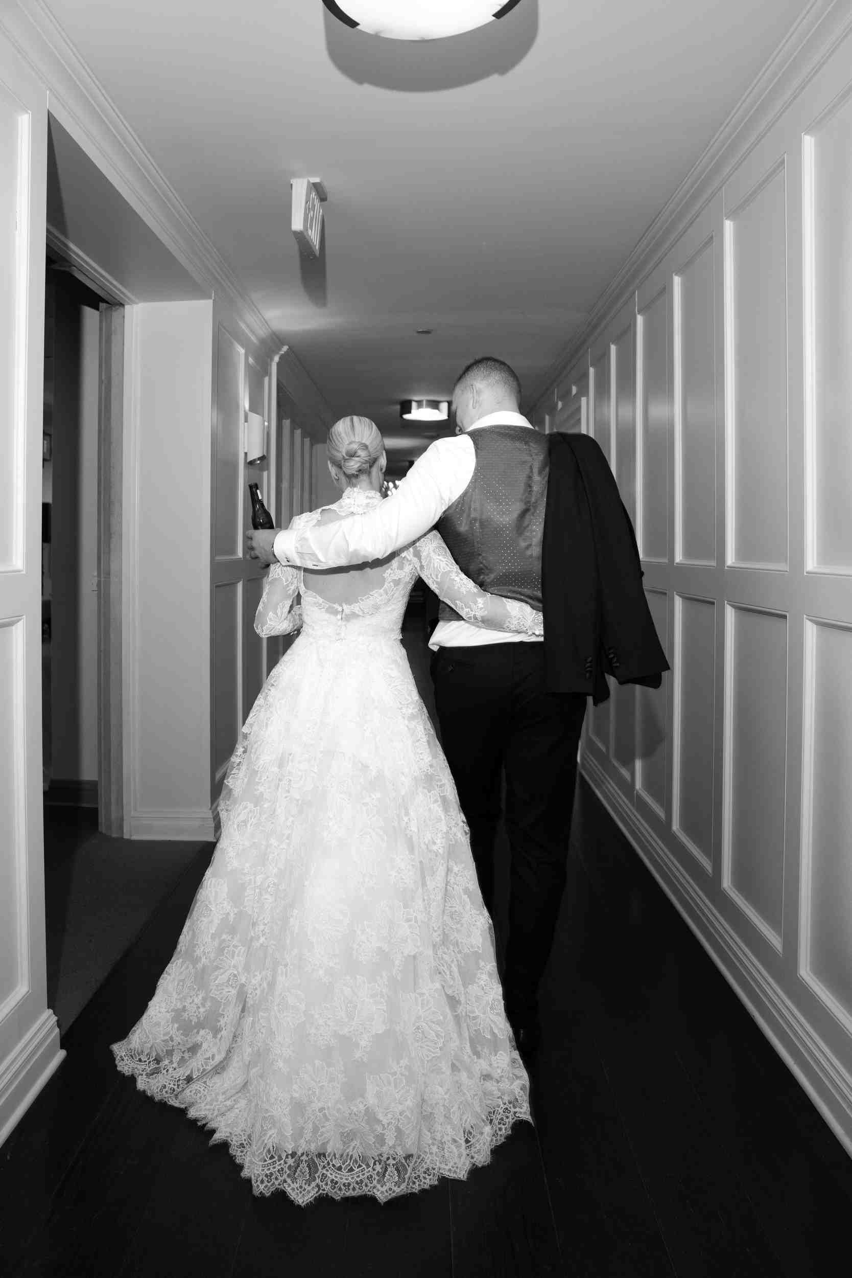 A bride and groom walking down a hall with wall paneling. The bride is wearing a lace wedding dress and holding a beer, and the groom is wearing a vest and holding a jacket over his shoulder.