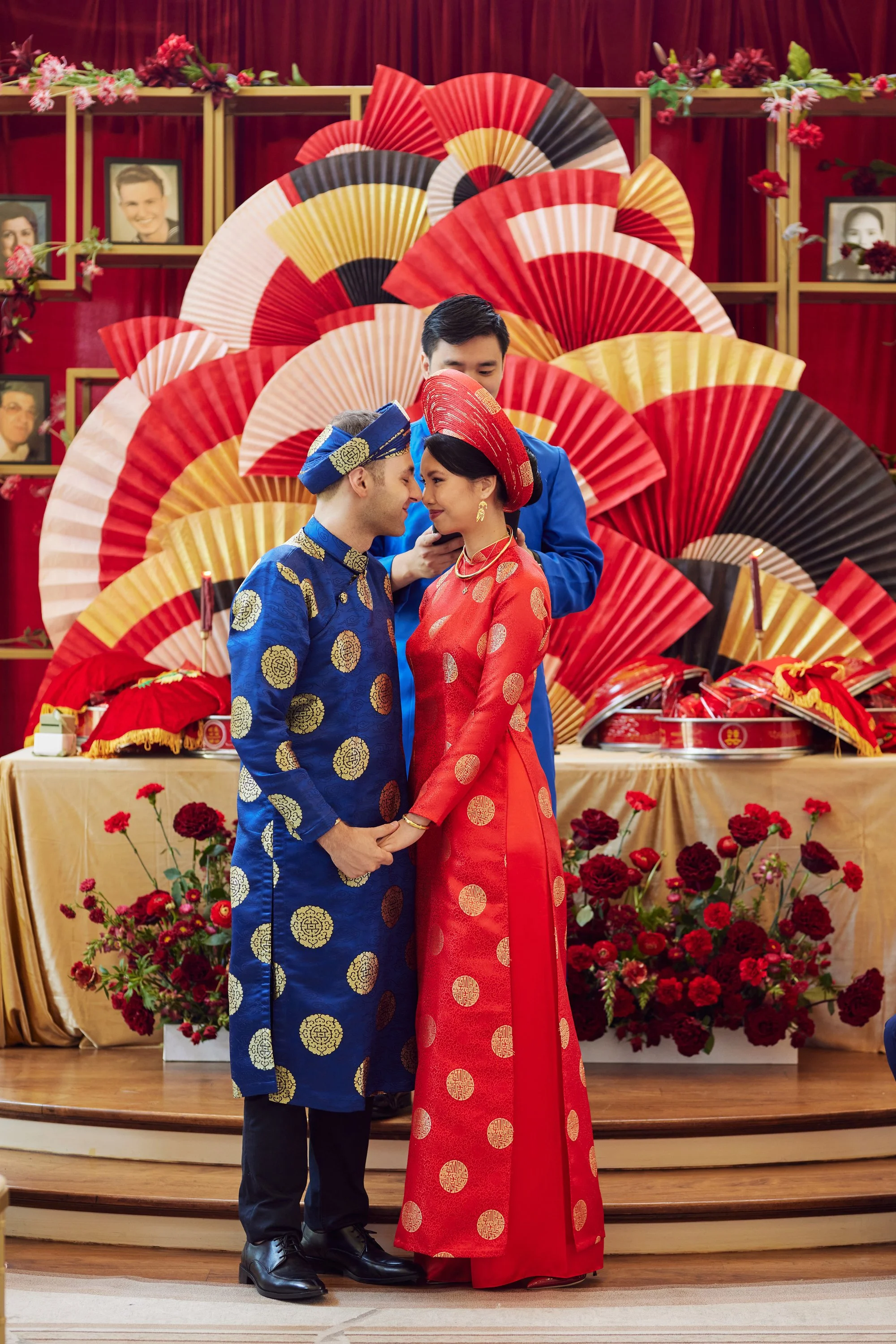A couple and a man at a traditional Vietnamese wedding, the couple dressed in áo dài; the groom in blue and the bride in red, with a backdrop of fans in red, yellow, white, and black, and framed photographs on a red background, decorated with flowers and candles.