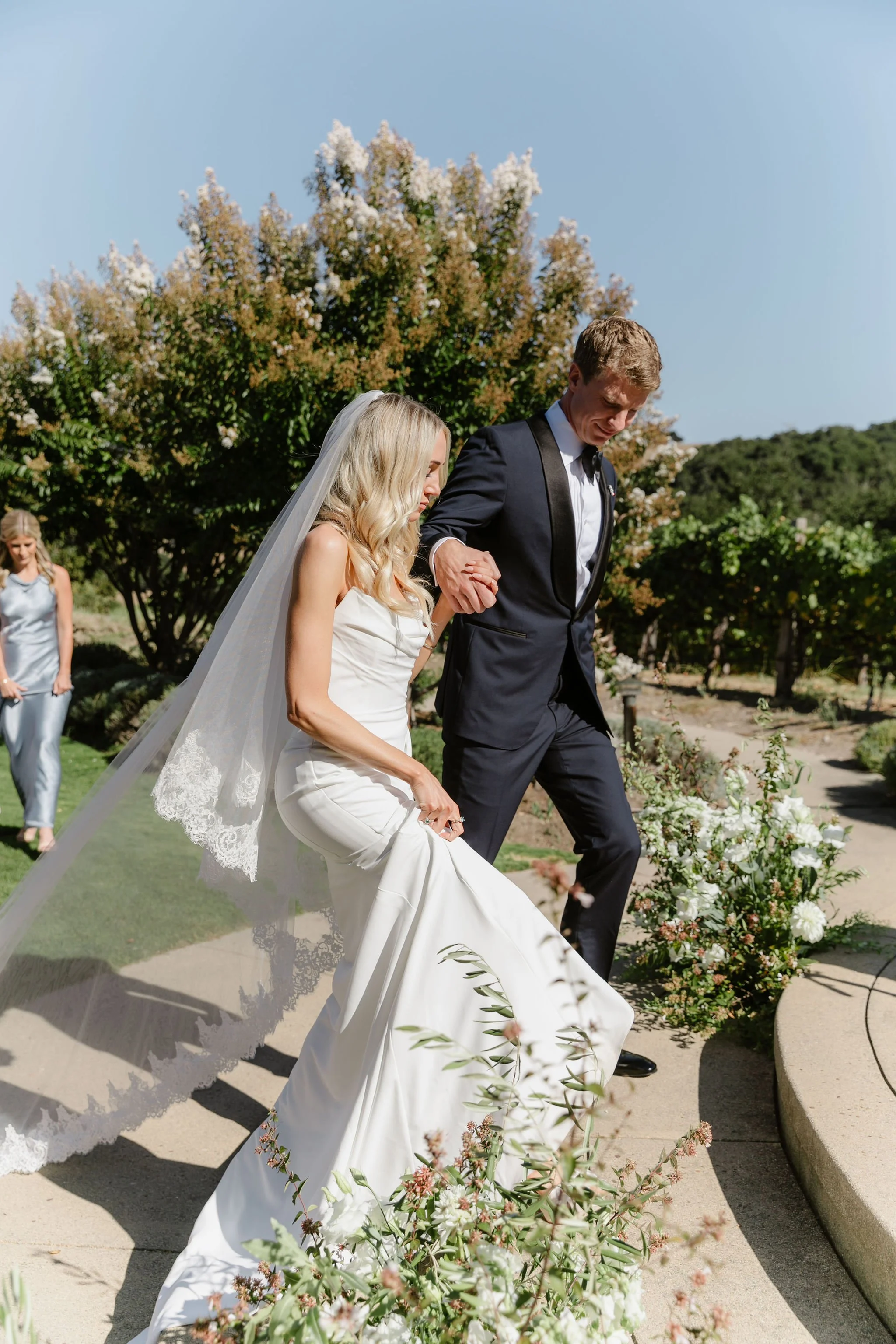 A bride and groom holding hands and walking outdoors during a wedding ceremony, with a bridesmaid in the background and floral arrangements along the path.
