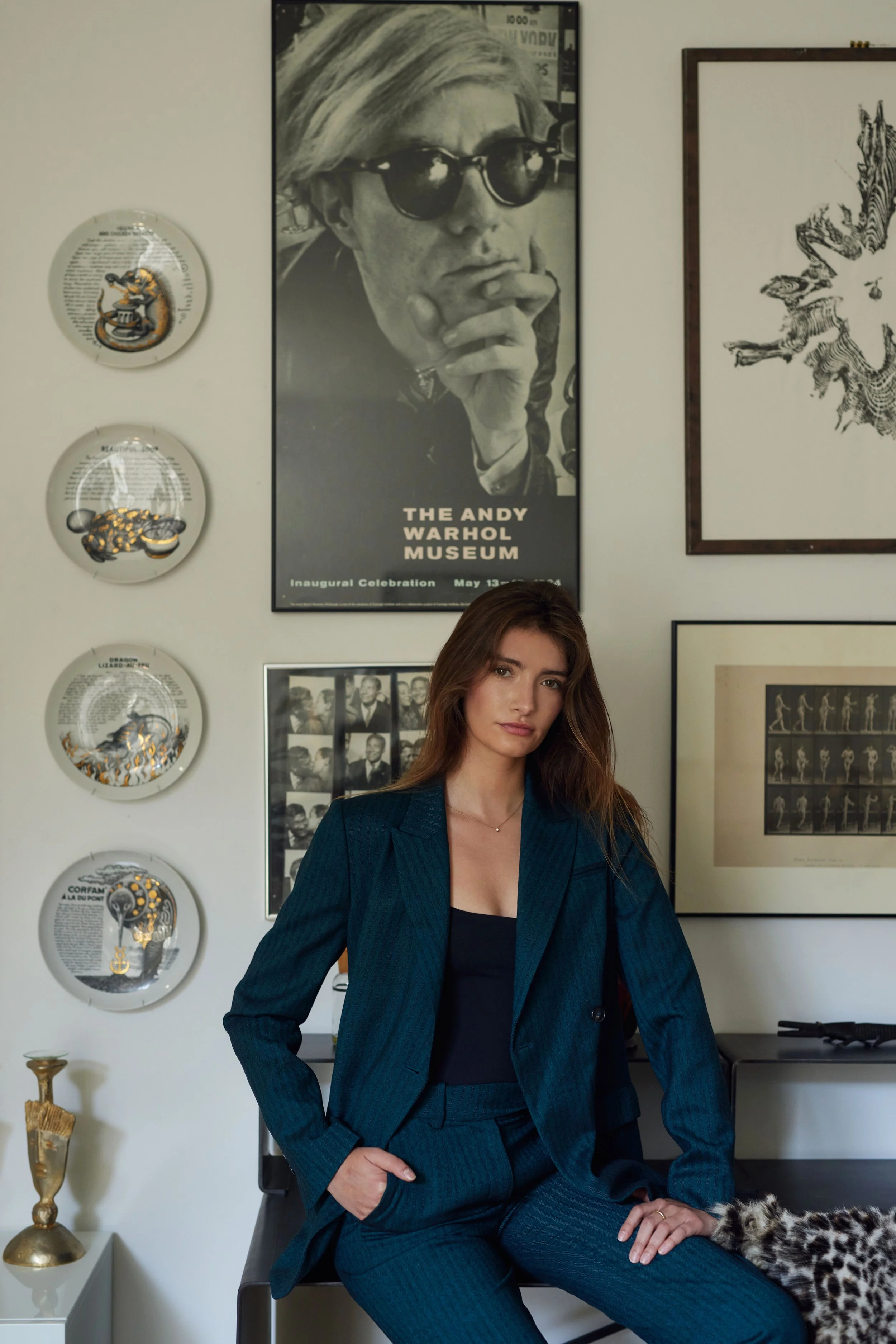 A woman with long brown hair and light skin wearing a dark teal pantsuit seated in a modern office or gallery space with framed artwork and decorative plates on the wall behind her.