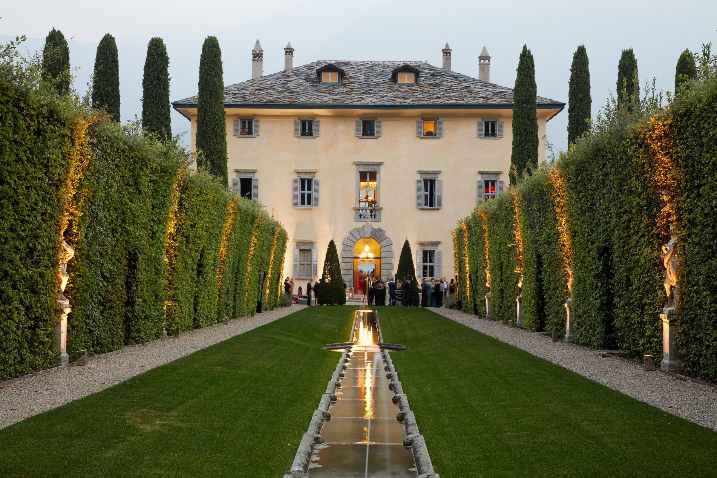 A large, elegant mansion with a stone roof, surrounded by tall, narrow trees. The mansion has multiple windows with shutters, some illuminated from inside. In the foreground, a symmetrical, manicured lawn with a narrow water feature running down the 