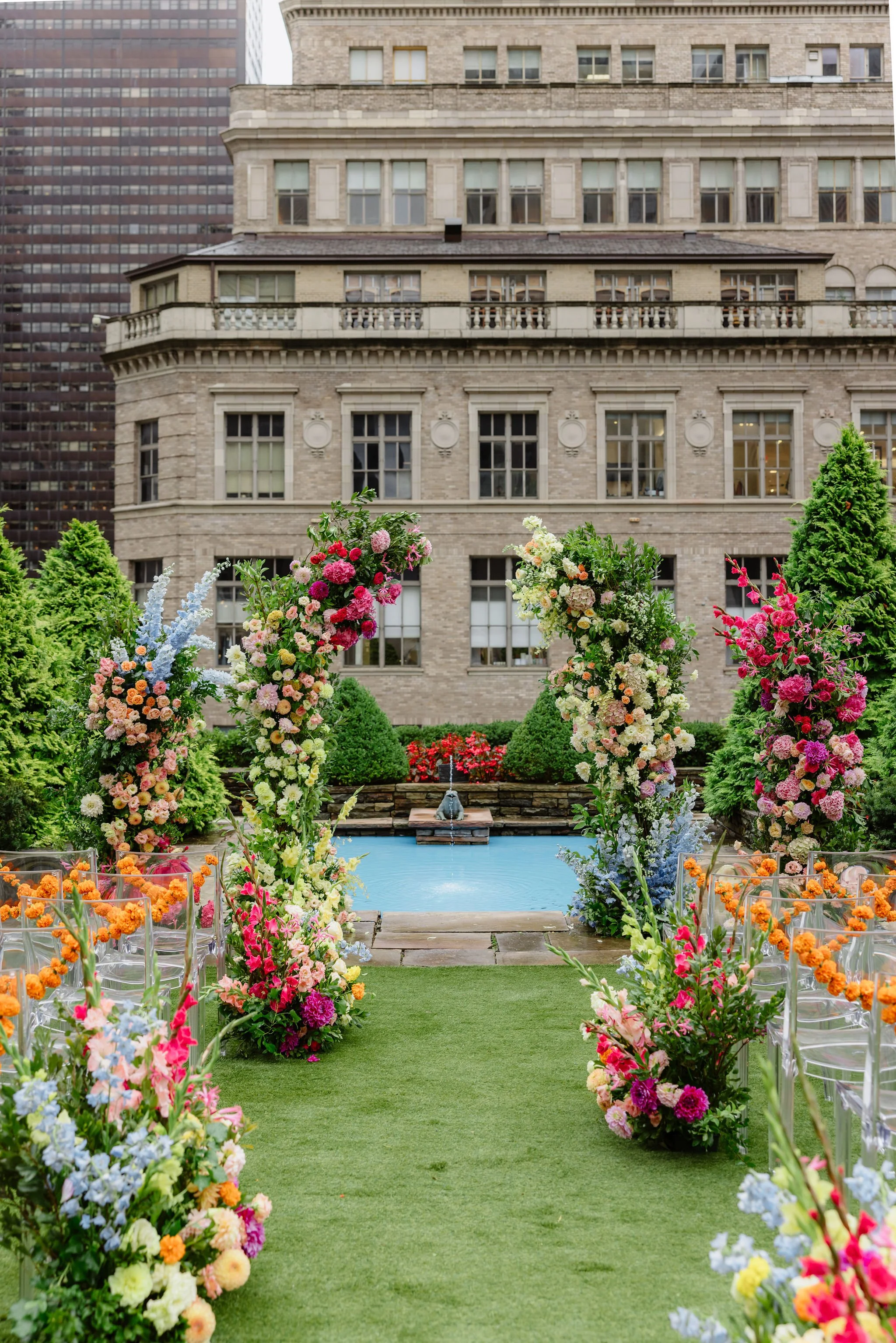A decorated outdoor aisle with colorful flowers on both sides leading to a pool, with a building and trees in the background.