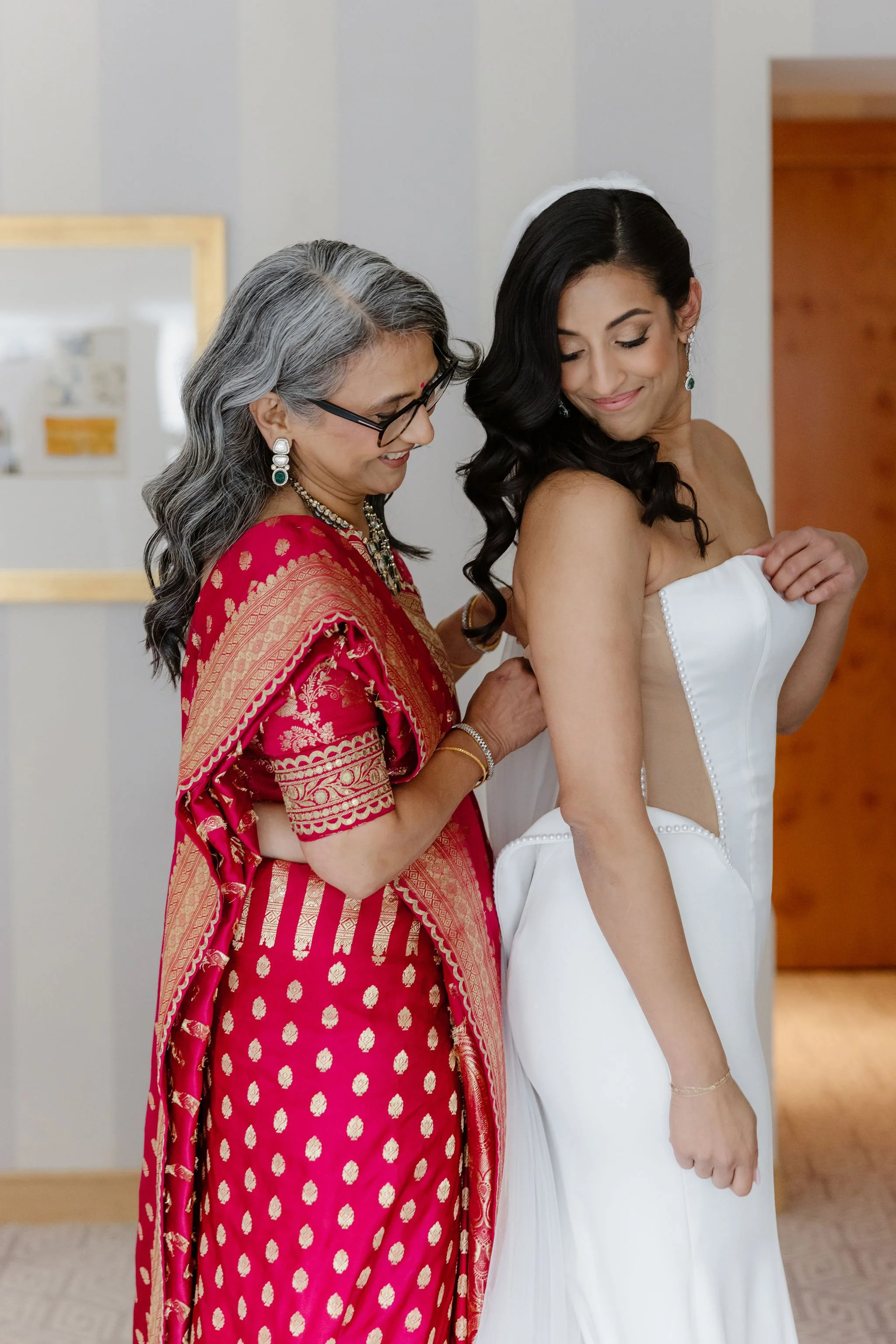 An older woman in a pink and gold traditional Indian saree helps a young woman in a white strapless dress with a corset back, likely preparing for a wedding or special occasion, in a room with light-colored walls and wooden accents.