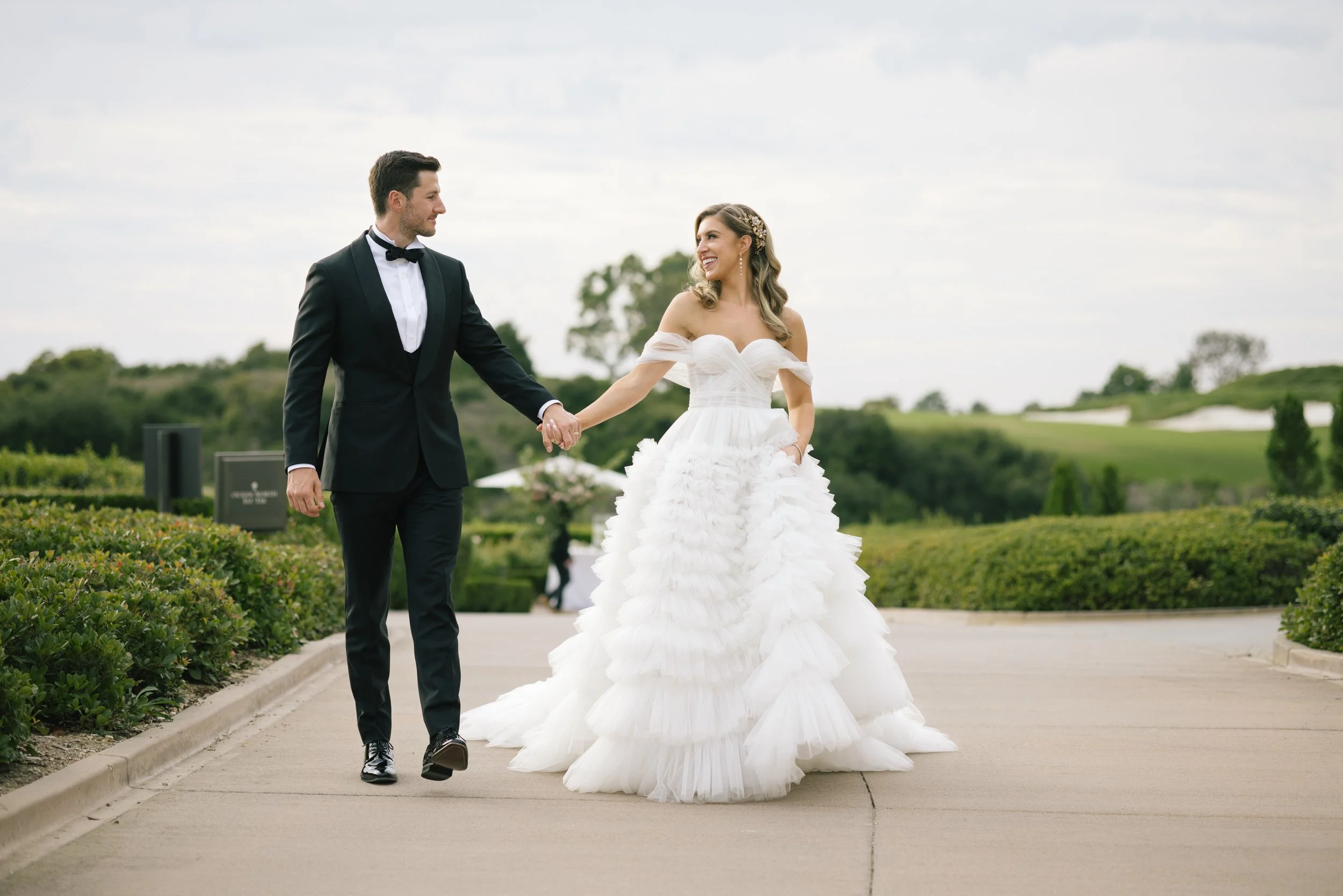 A bride and groom walking hand in hand outdoors on a cloudy day. The groom is dressed in a black tuxedo with a bow tie, and the bride is wearing a white off-the-shoulder gown with ruffled layers. They are smiling at each other, surrounded by greenery