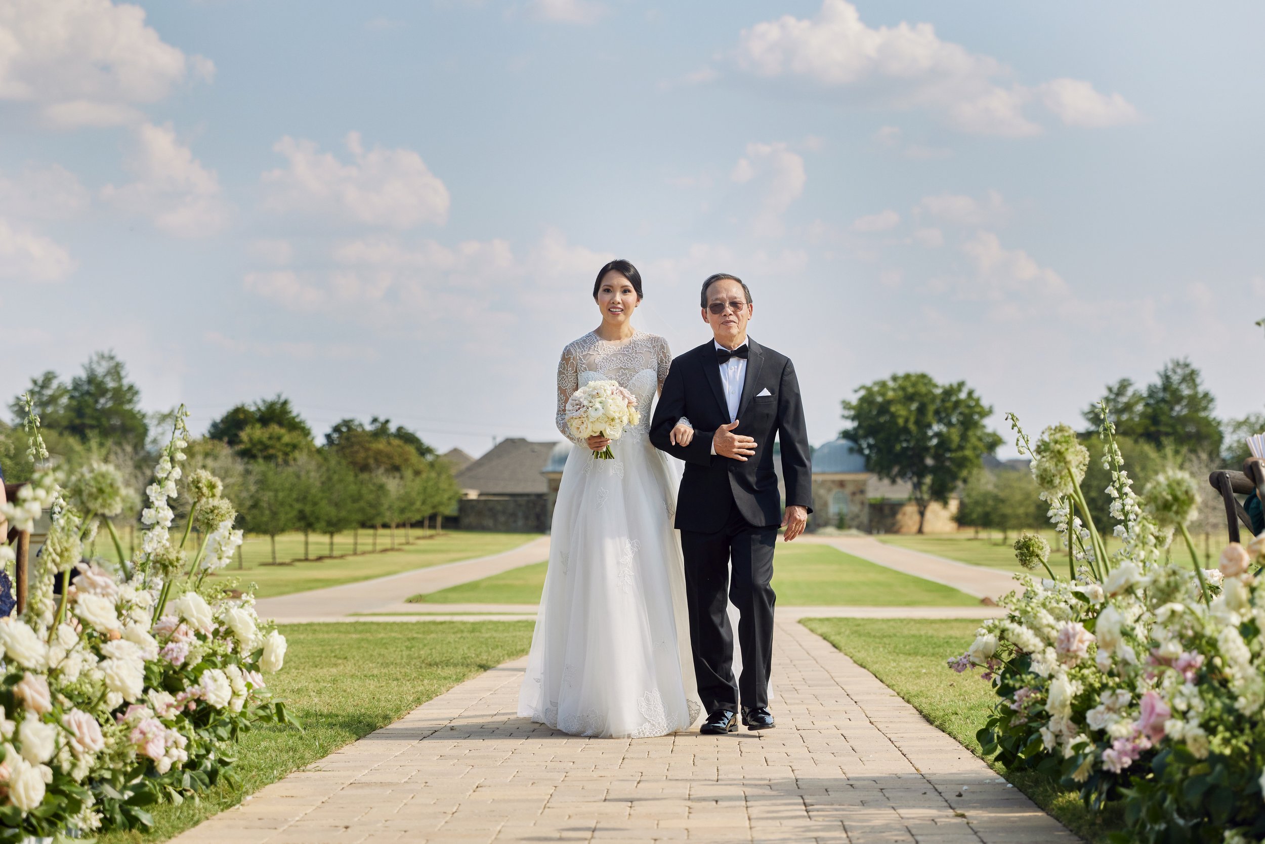 A bride in a white lace wedding dress holding a bouquet, walking down an outdoor aisle with a man in a tuxedo. The aisle is decorated with white flowers, and there are trees and houses in the background under a partly cloudy sky.