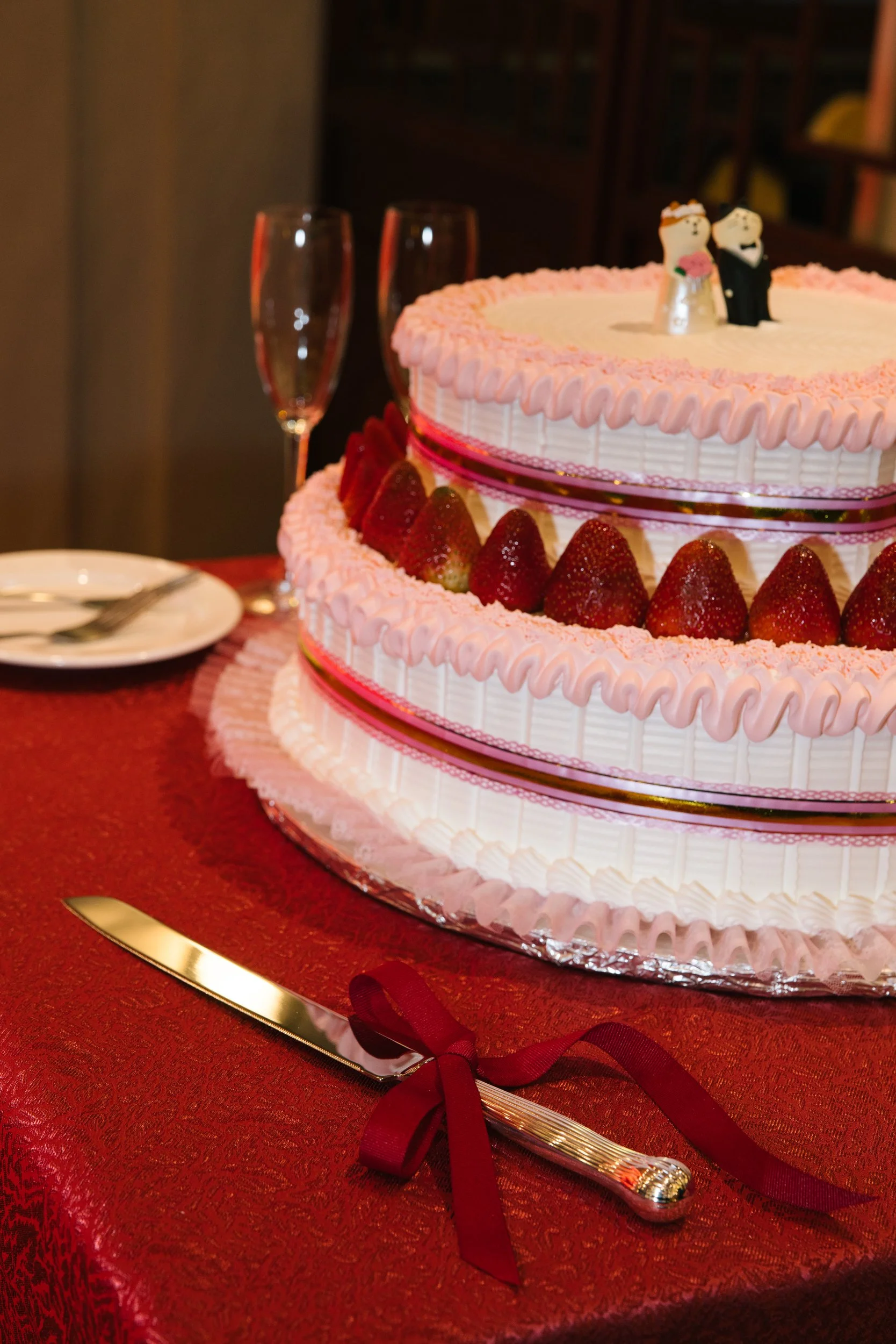 A tiered wedding cake decorated with pink and white frosting, strawberries, and bride and groom figurines. The cake is on a table with a red tablecloth, flanked by two empty champagne glasses, a plate with a fork, and a gold-colored knife with a red 