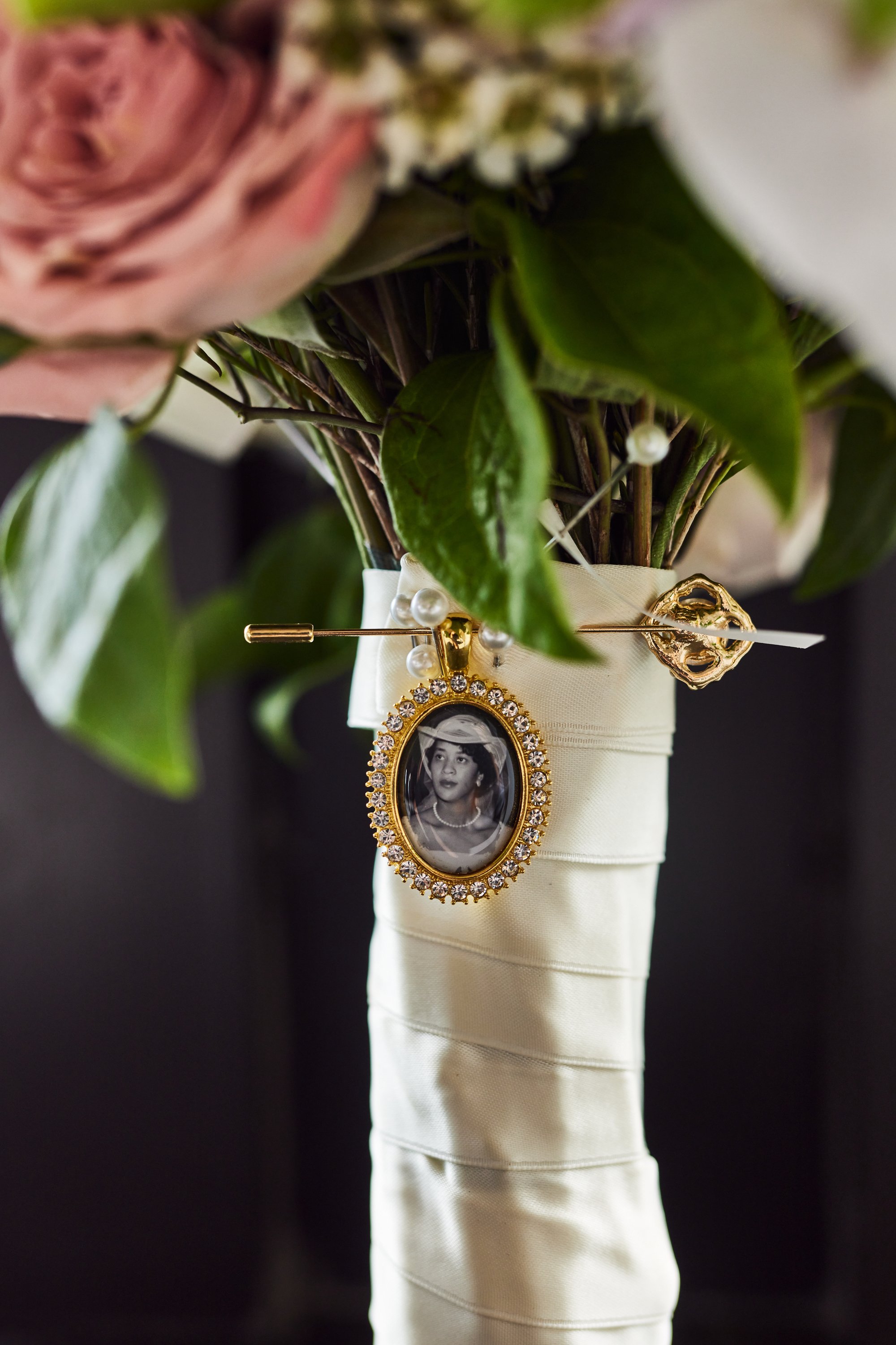 Close-up of a bouquet of pink and white roses with green leaves, wrapped in white satin fabric, with a cameo brooch featuring a black-and-white portrait of a woman and gold decorative elements, against a dark background.