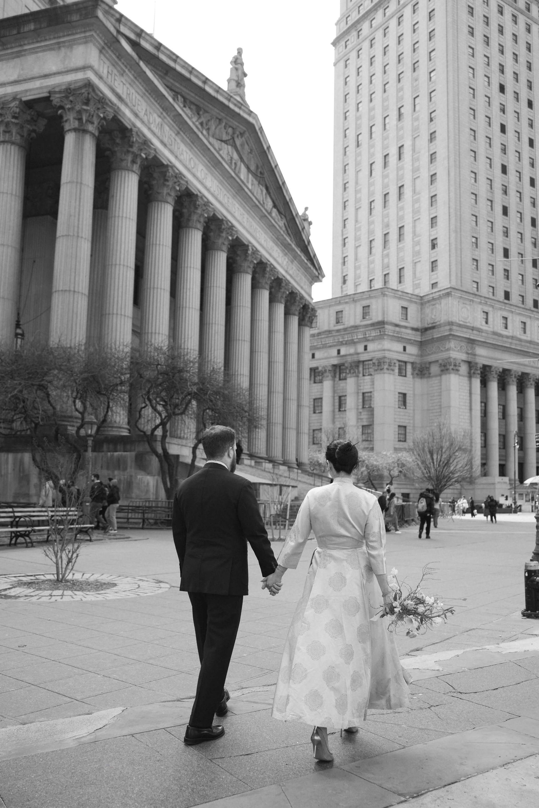 A black-and-white photo of a man and woman holding hands, walking in an urban park. The woman is wearing a long dress with large floral patterns and high heels, carrying a bouquet of flowers. The man is dressed in a suit. Tall buildings and a classic