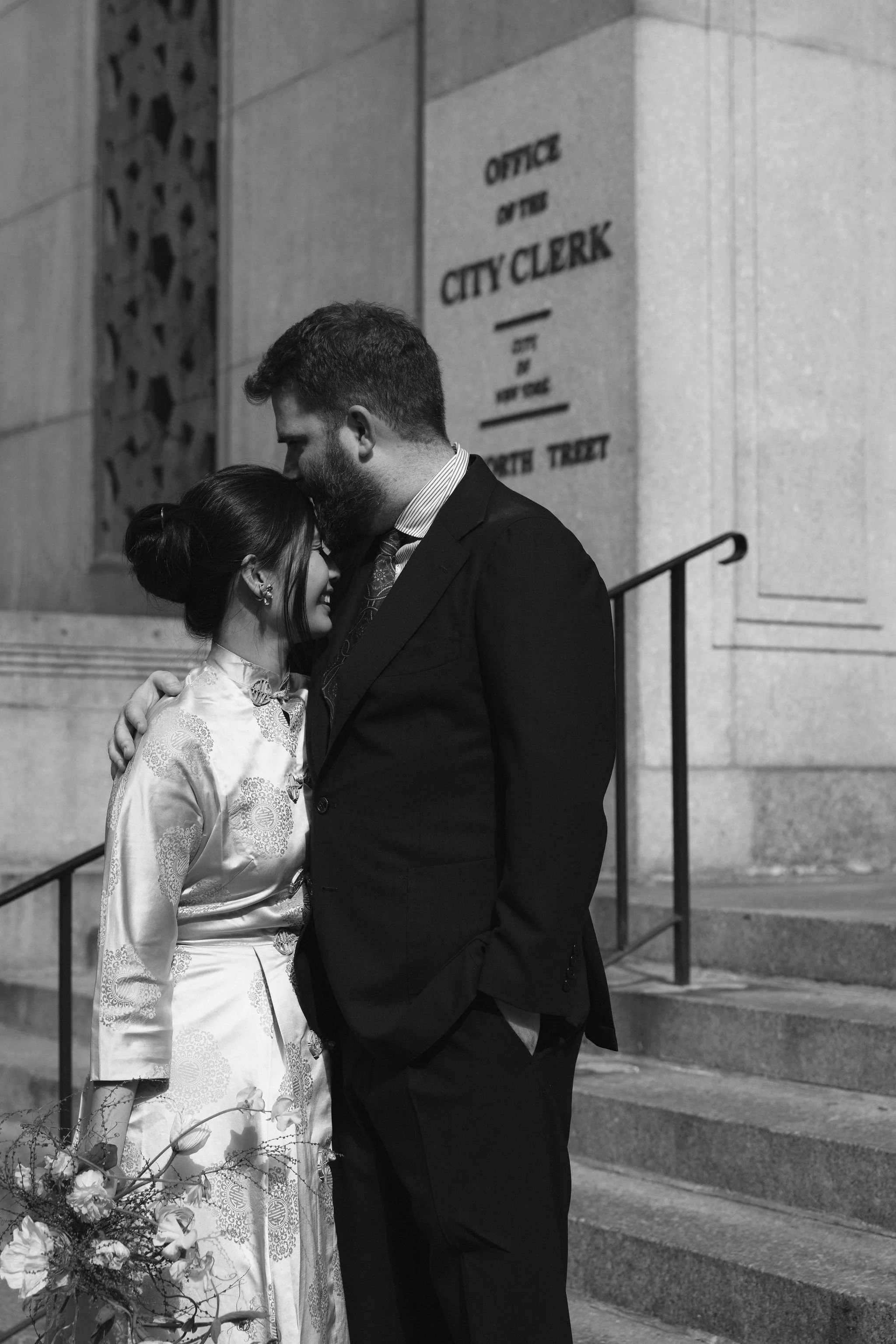 A black and white photo of a couple embracing outside a city hall, with a sign that says "Office of the City Clerk" on the building behind them.