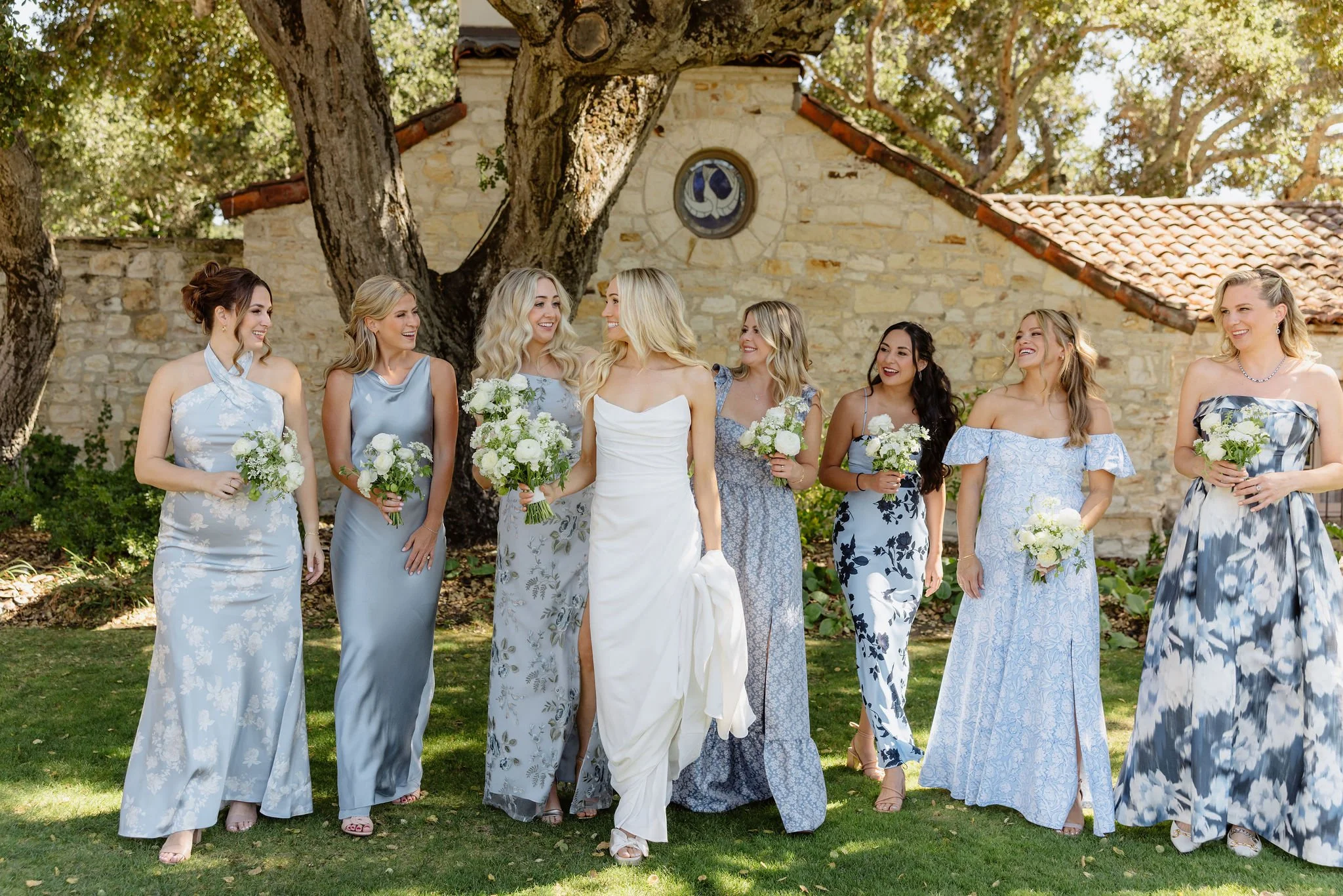 A group of women dressed in blue and white floral dresses holding white flower bouquets, standing outdoors in front of a stone building and large trees, during a wedding celebration.
