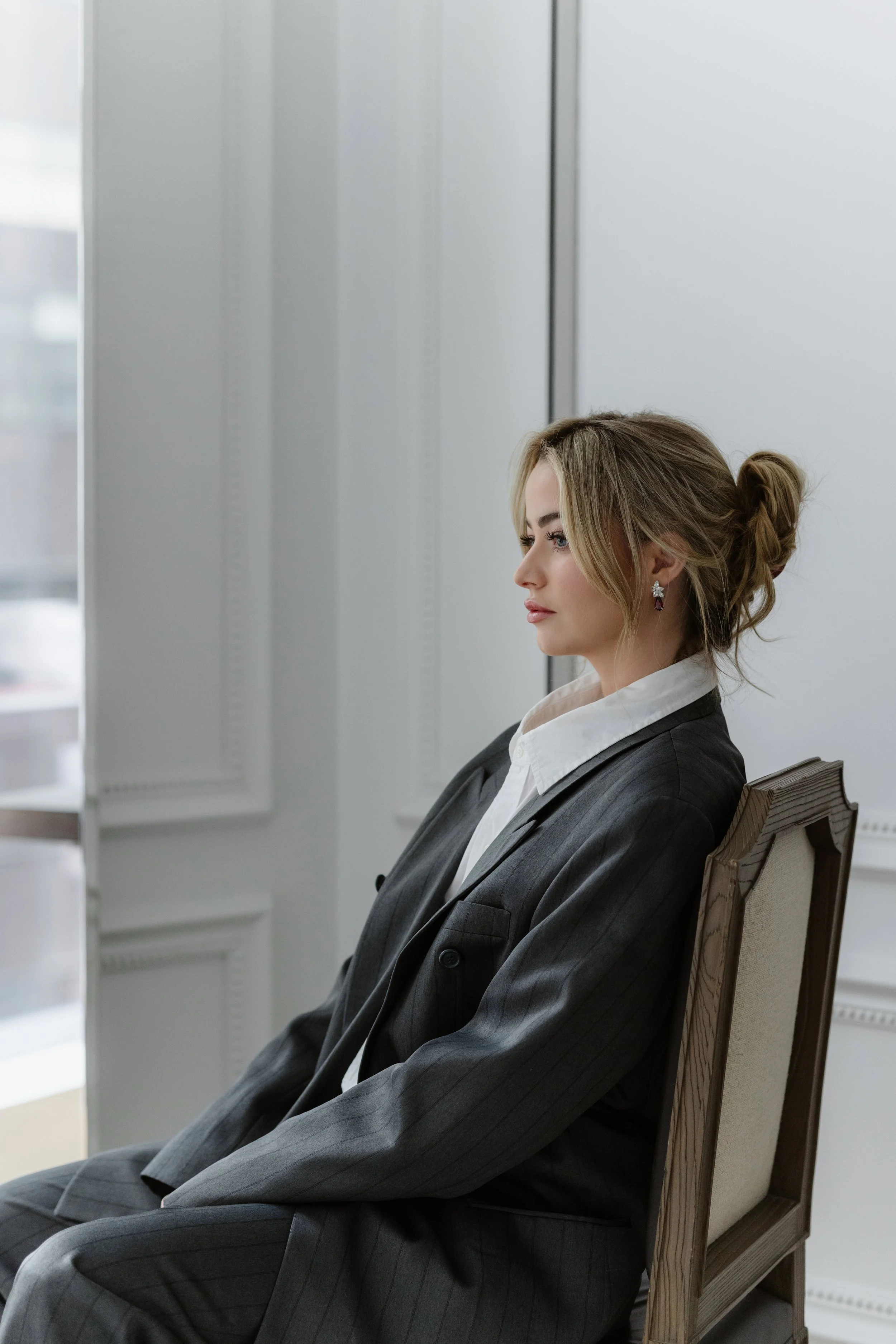 A woman with blonde hair styled in a loose bun, dressed in a dark gray pinstripe suit and white shirt, sitting on a wooden chair with a cream-colored cushion in a room with white walls and a large window.