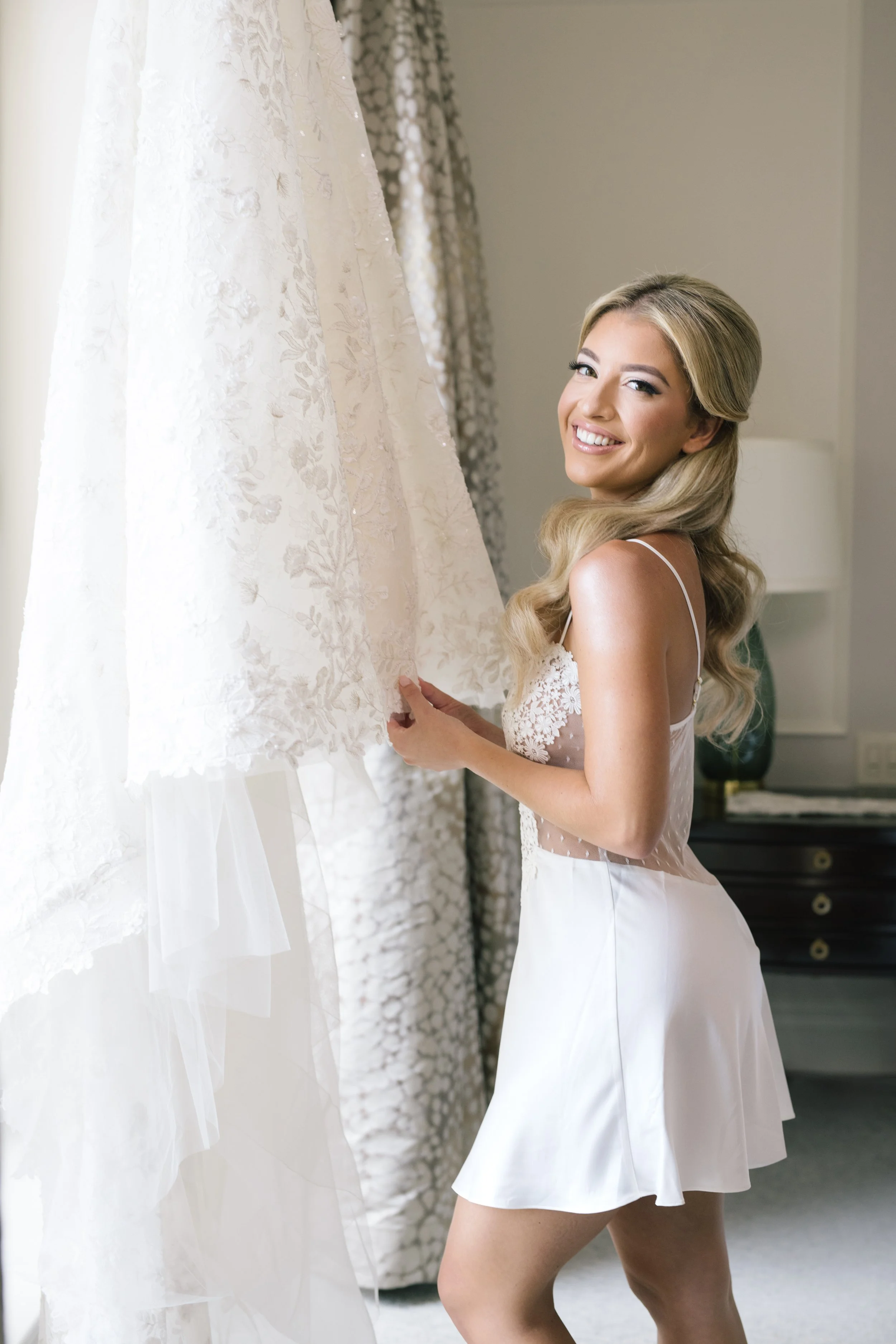 A smiling woman with long, wavy blonde hair and makeup is wearing a white lingerie slip and standing next to a wedding dress with lace detail, holding and looking at it in a room with patterned curtains.