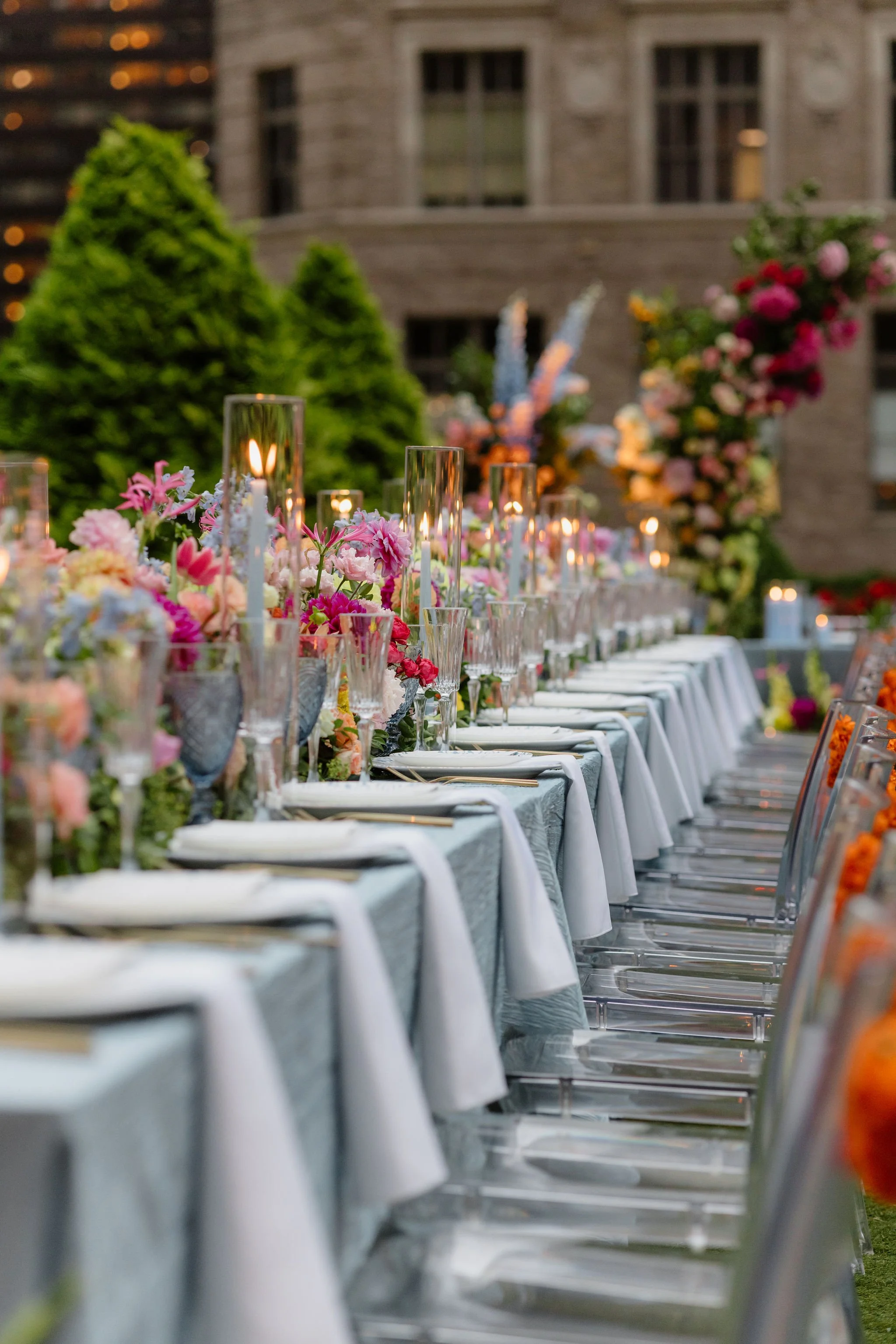 Long outdoor table set for a wedding or event, decorated with colorful flowers in glass vases and candle lanterns, with greenery and urban buildings in the background.