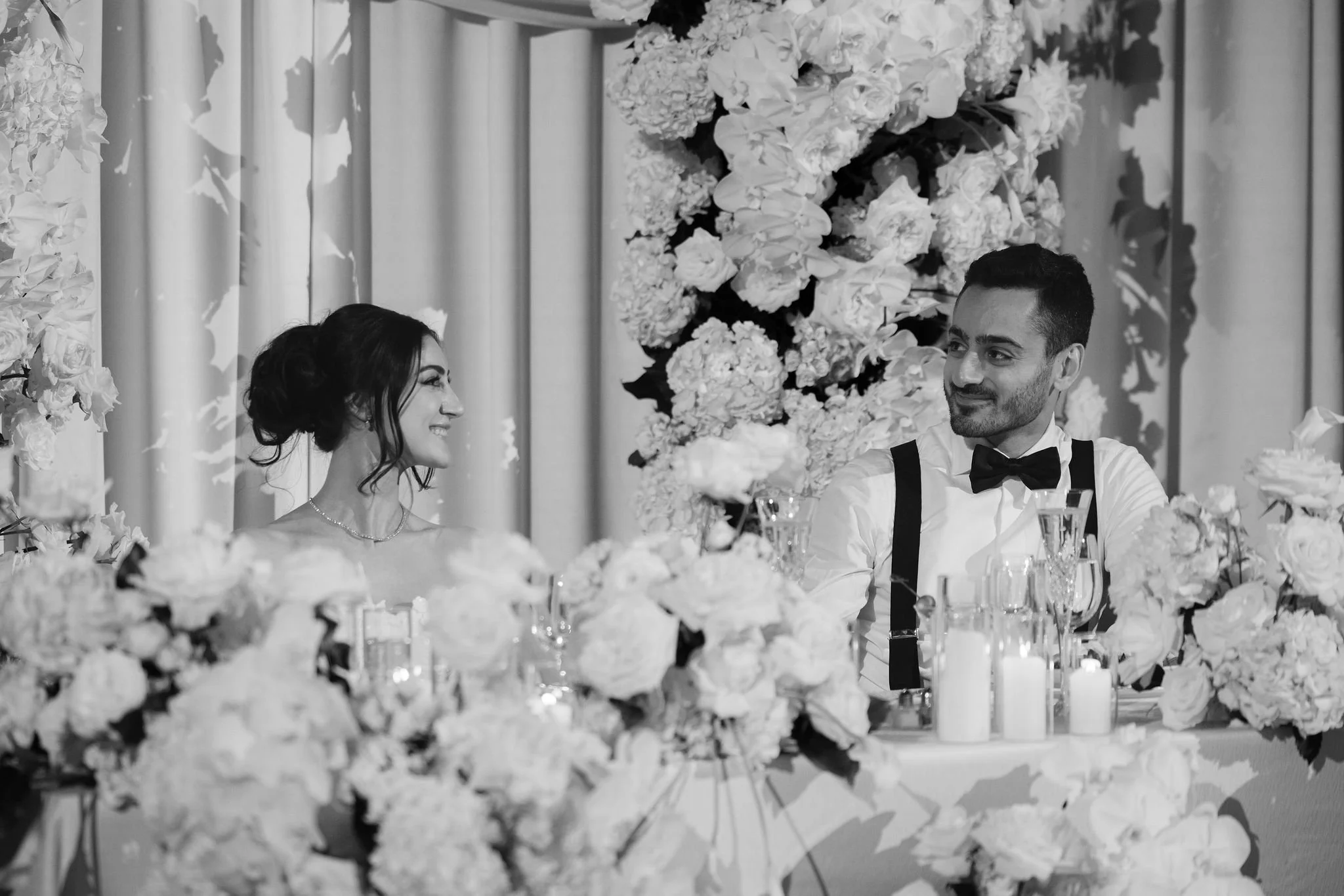 A black and white photo of a bride and groom at their wedding reception, sitting at a table decorated with flowers and candles. The bride is smiling at the groom, and the groom is looking at her.