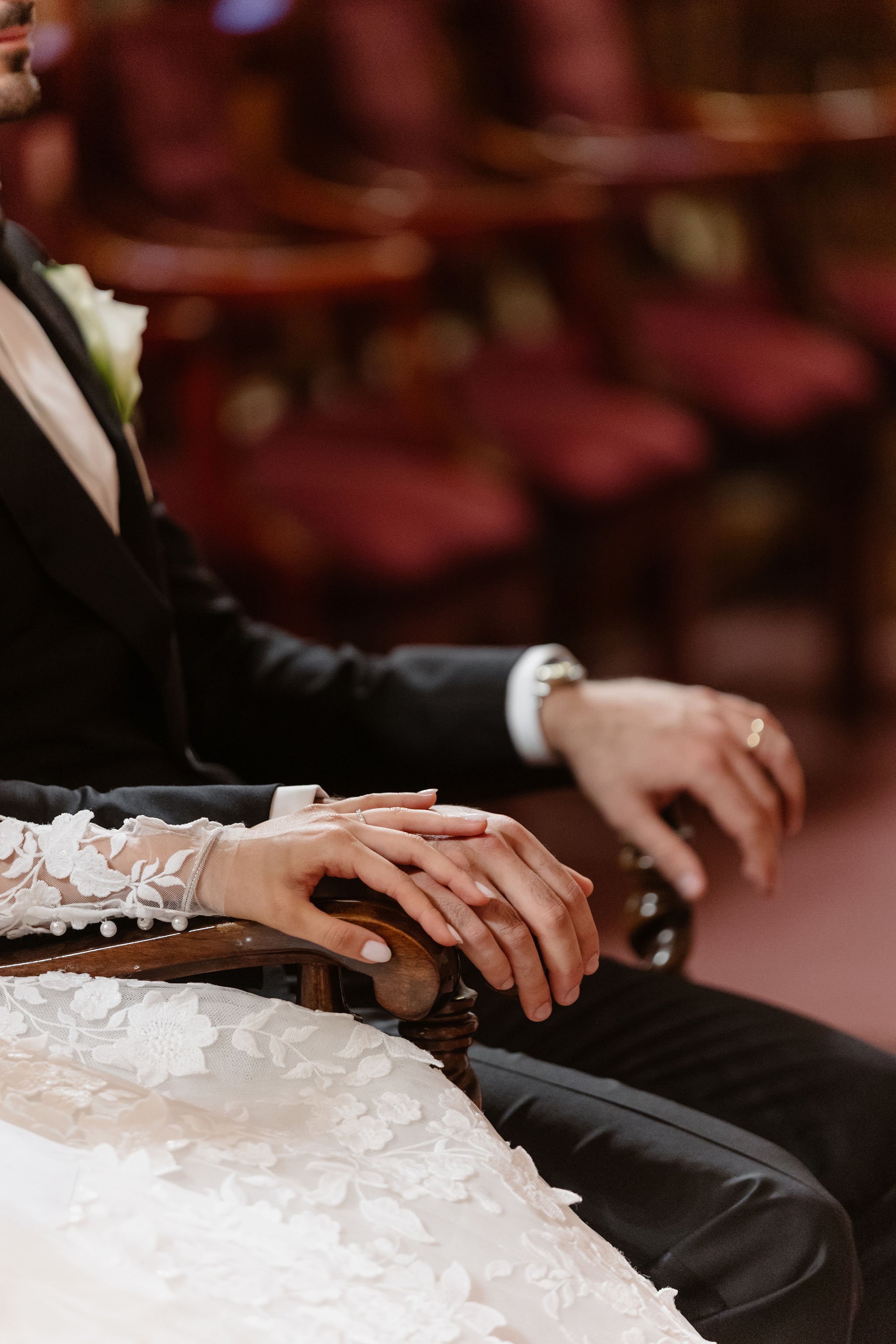 Close-up of wedding couple's hands resting on a wooden armrest, with the bride's hand adorned with a ring and lace sleeve, and the groom's hand next to hers, both wearing formal attire.