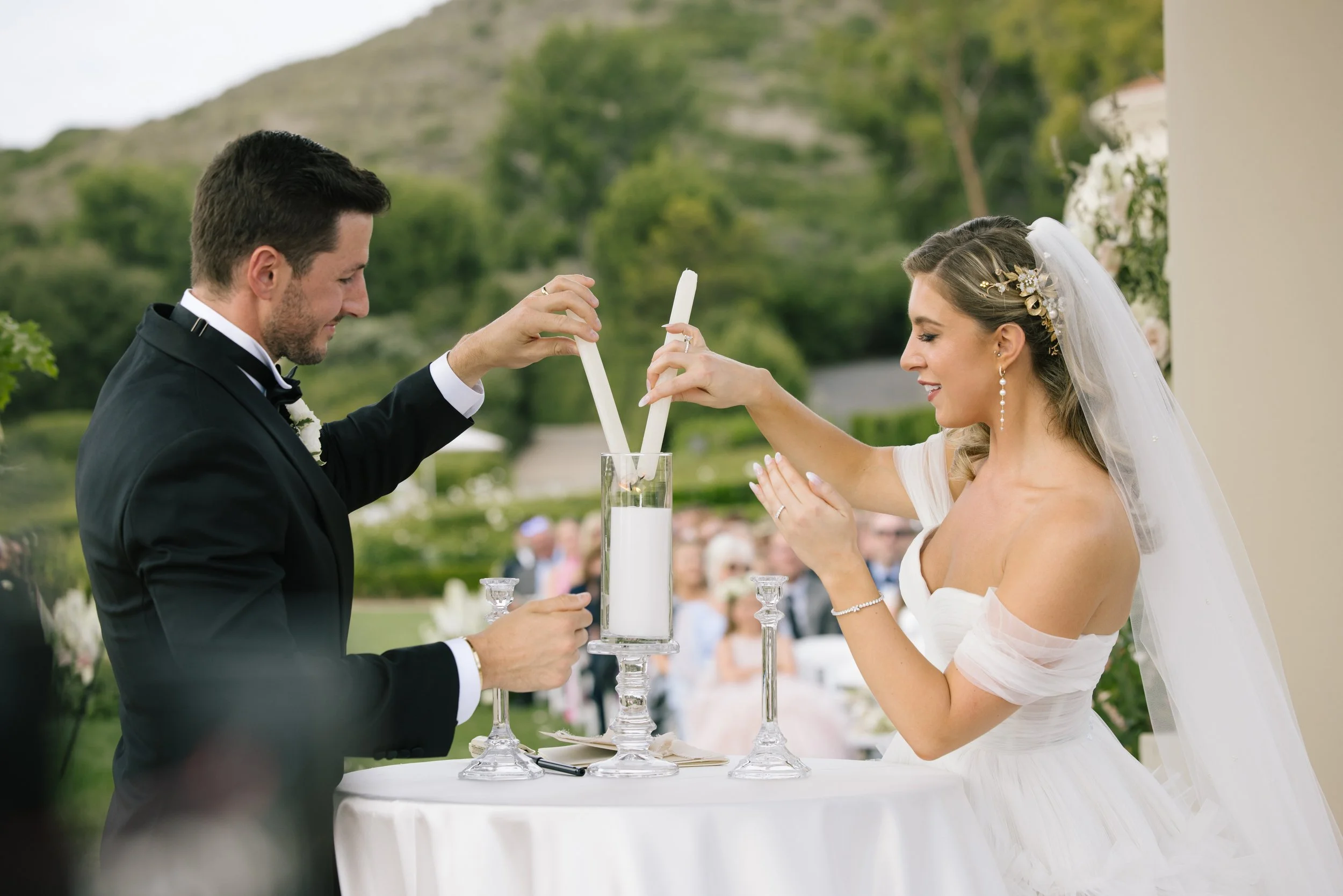 Bride and groom exchanging candles during wedding ceremony outdoors with greenery and mountain in background.