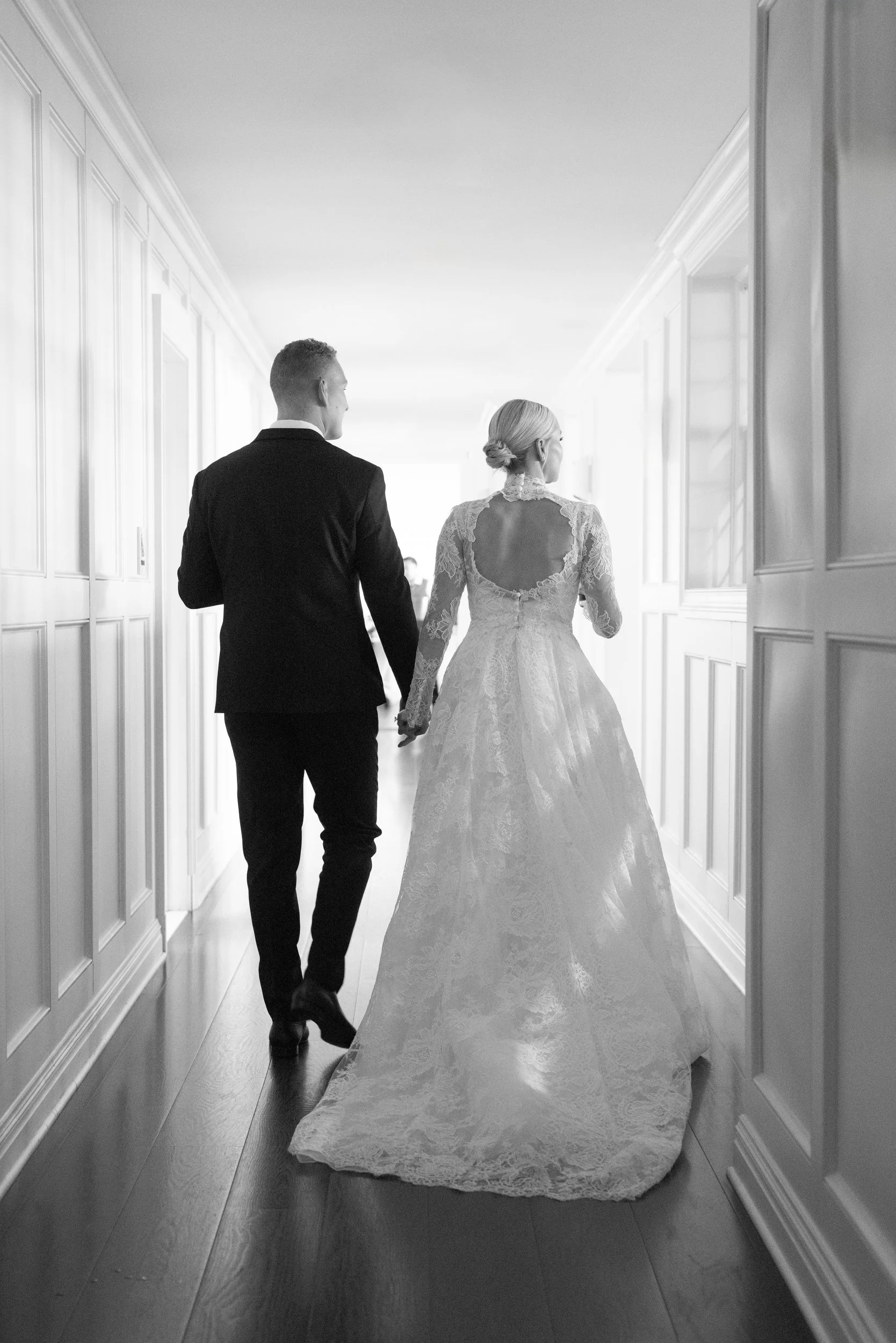 A bride and groom walk hand in hand down a hallway, with the bride in a lace wedding gown and the groom in a black suit.