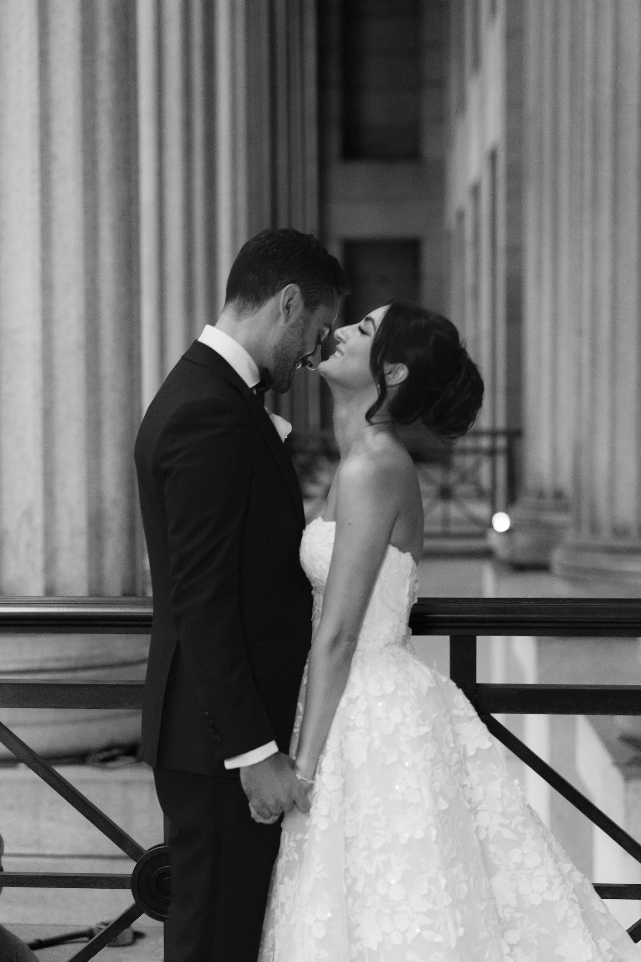 A bride and groom dressed in wedding attire sharing a moment in front of a grand, classical interior with tall columns and curtains.