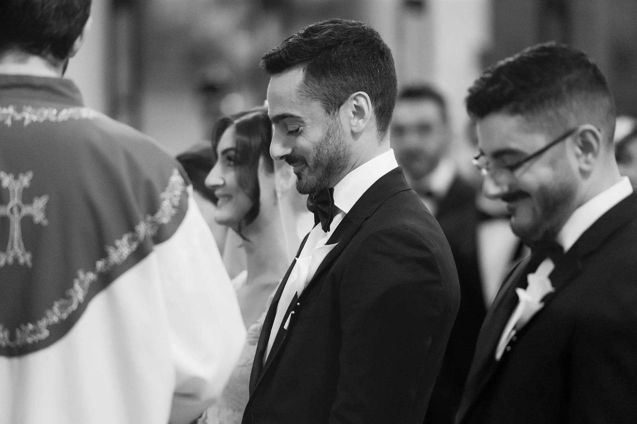 Black and white photo of a wedding ceremony with a groom in a tuxedo and bow tie smiling and standing with a line of people, some of whom are wearing glasses and formal attire, in a setting that appears to be a church or hall.