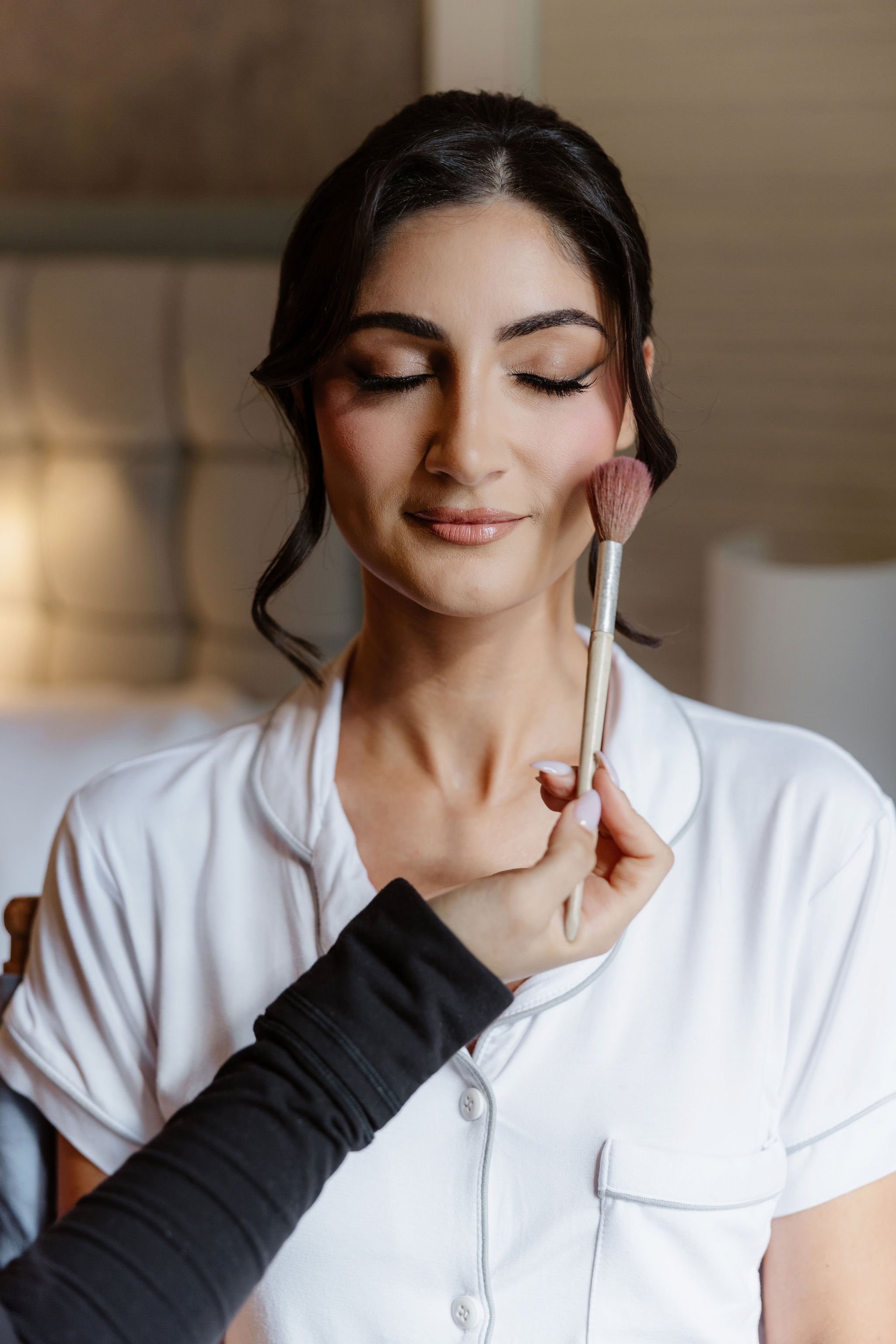 A woman with dark hair and makeup has her face gently touched with a makeup brush.
