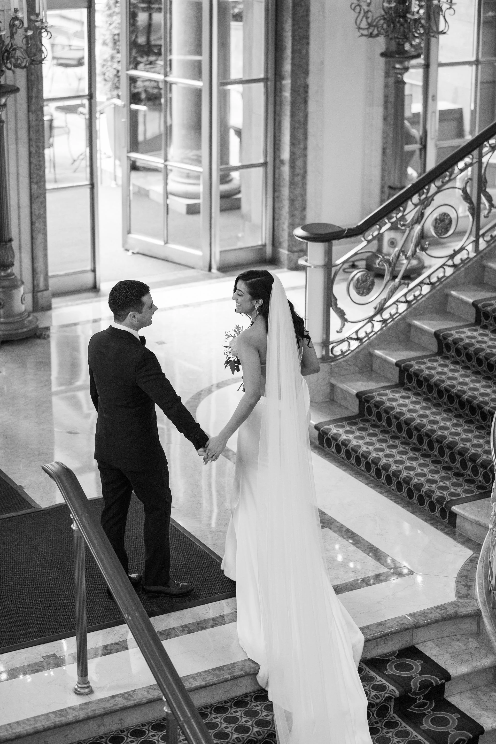 A black and white photo of a bride and groom holding hands on a staircase inside a building, with a large window and decorative staircase railing in the background.