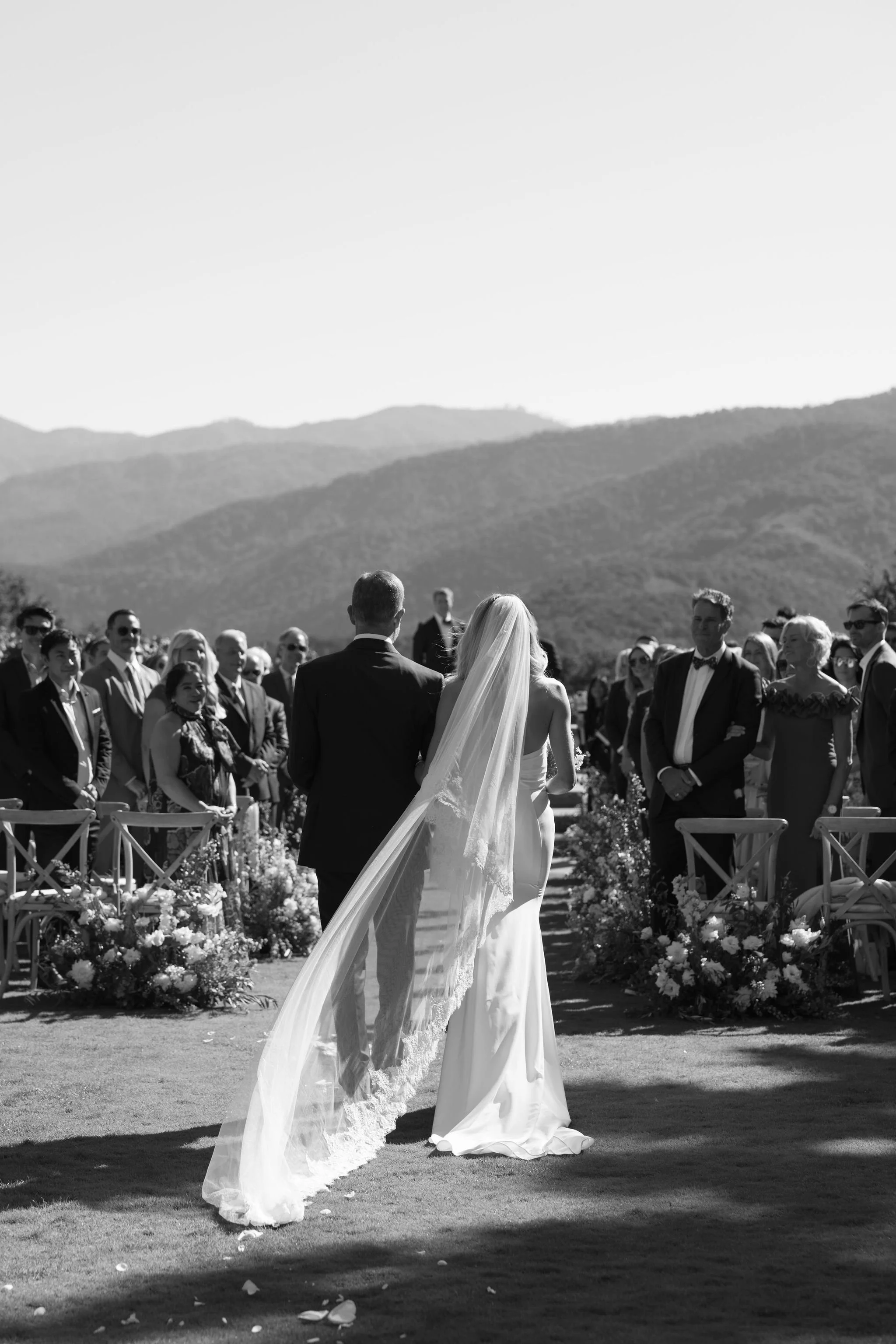 A black and white photo of a bride and groom at an outdoor wedding ceremony, with guests in the background, mountains, and a clear sky.