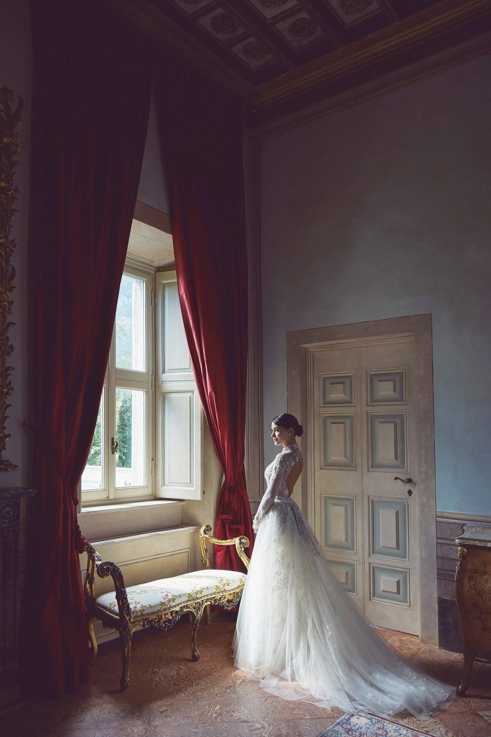 A woman in a white wedding dress standing near a window with red curtains in an elegant, vintage-style room with detailed moldings and furniture.