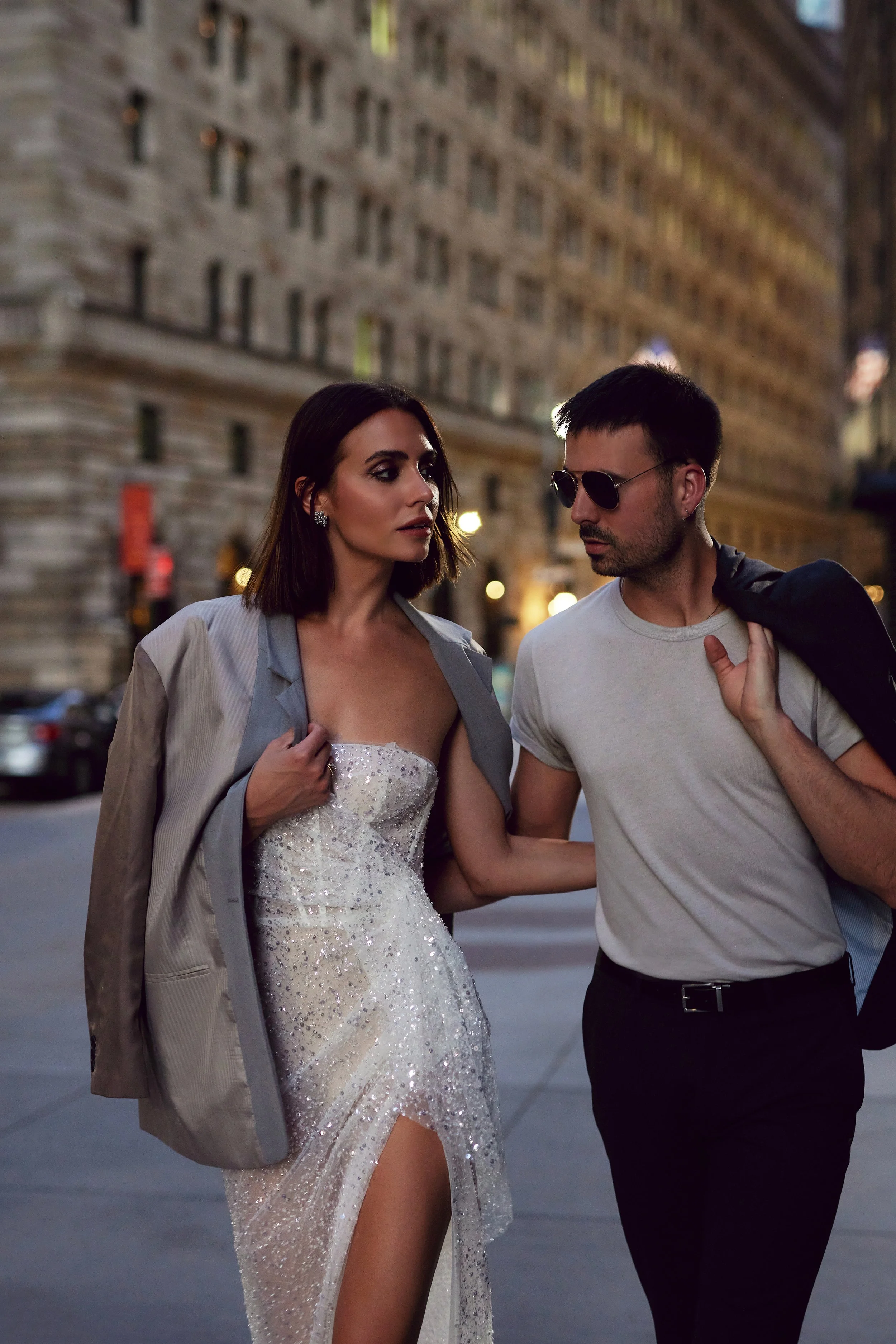 A woman in a sparkling white dress with a thigh-high slit and a man in a white t-shirt and sunglasses walking together in an urban setting at night. The woman is adjusting her dress and the man is carrying a jacket over his shoulder, looking at her.