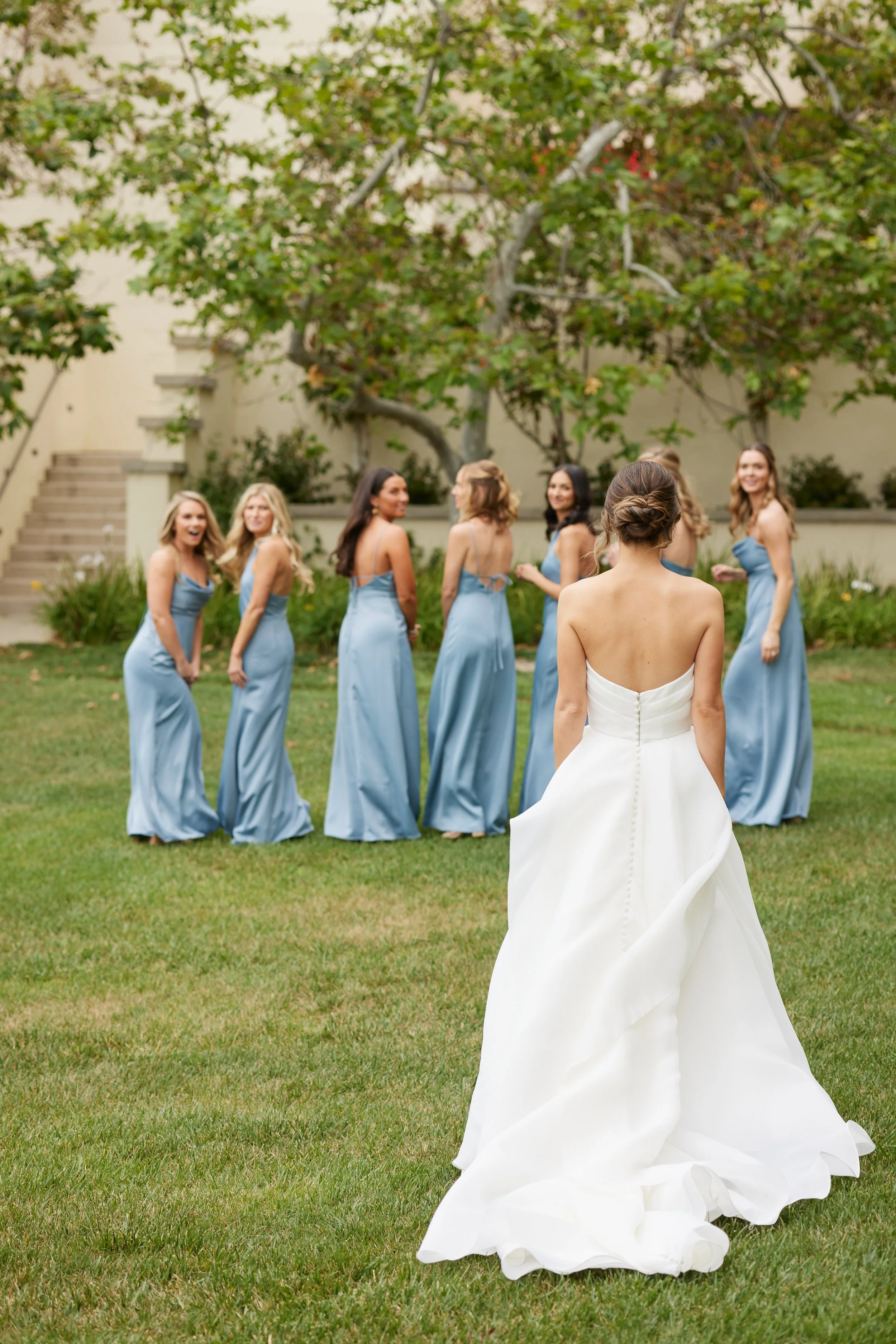 A bride in a white wedding dress standing outdoors on grass, facing a group of women in light blue bridesmaid dresses, with trees and a staircase in the background.