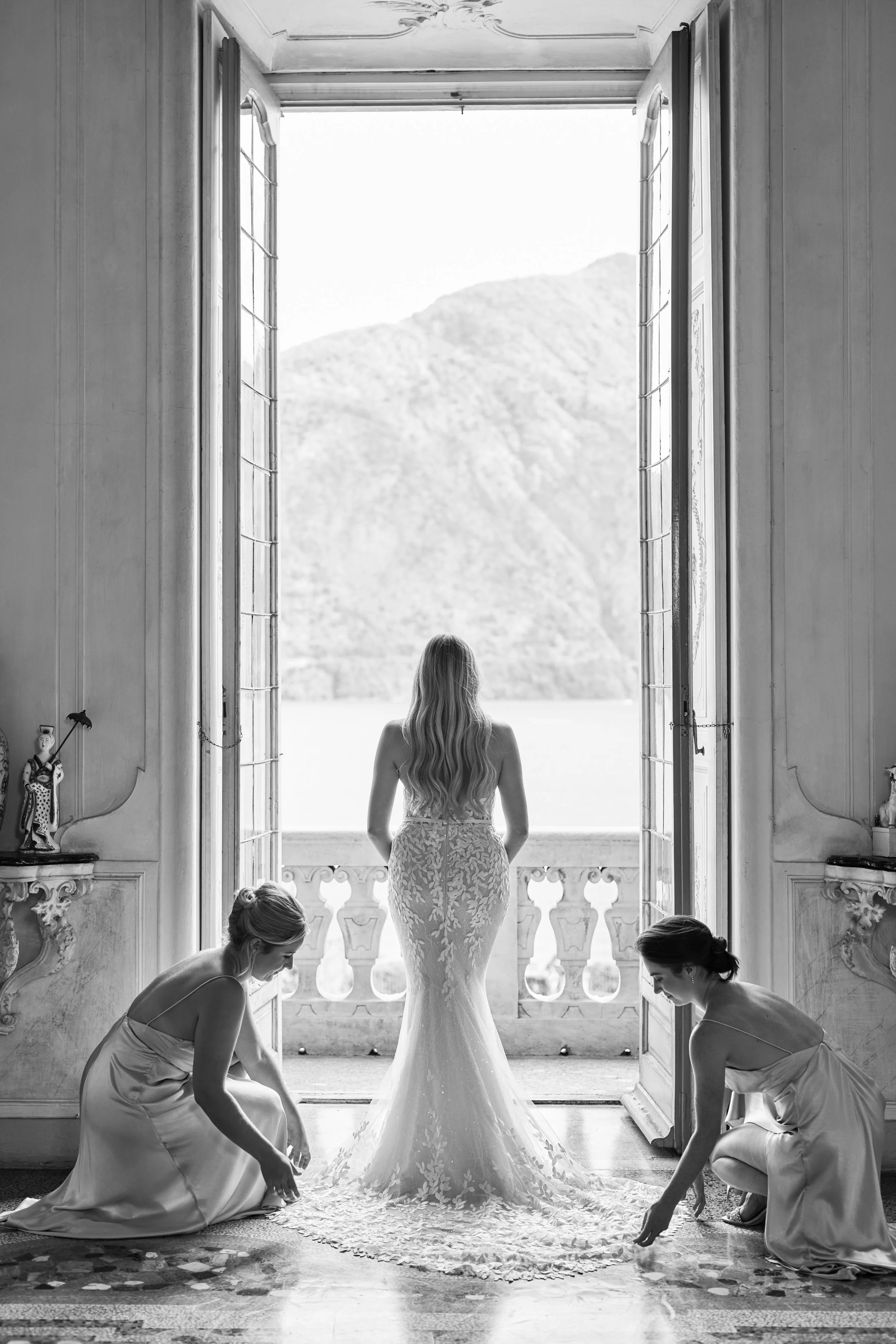 A bride in a wedding dress standing in a doorway with three women kneeling and working on the train of her dress, inside a room with ornate decor and an open balcony door revealing mountains outside.