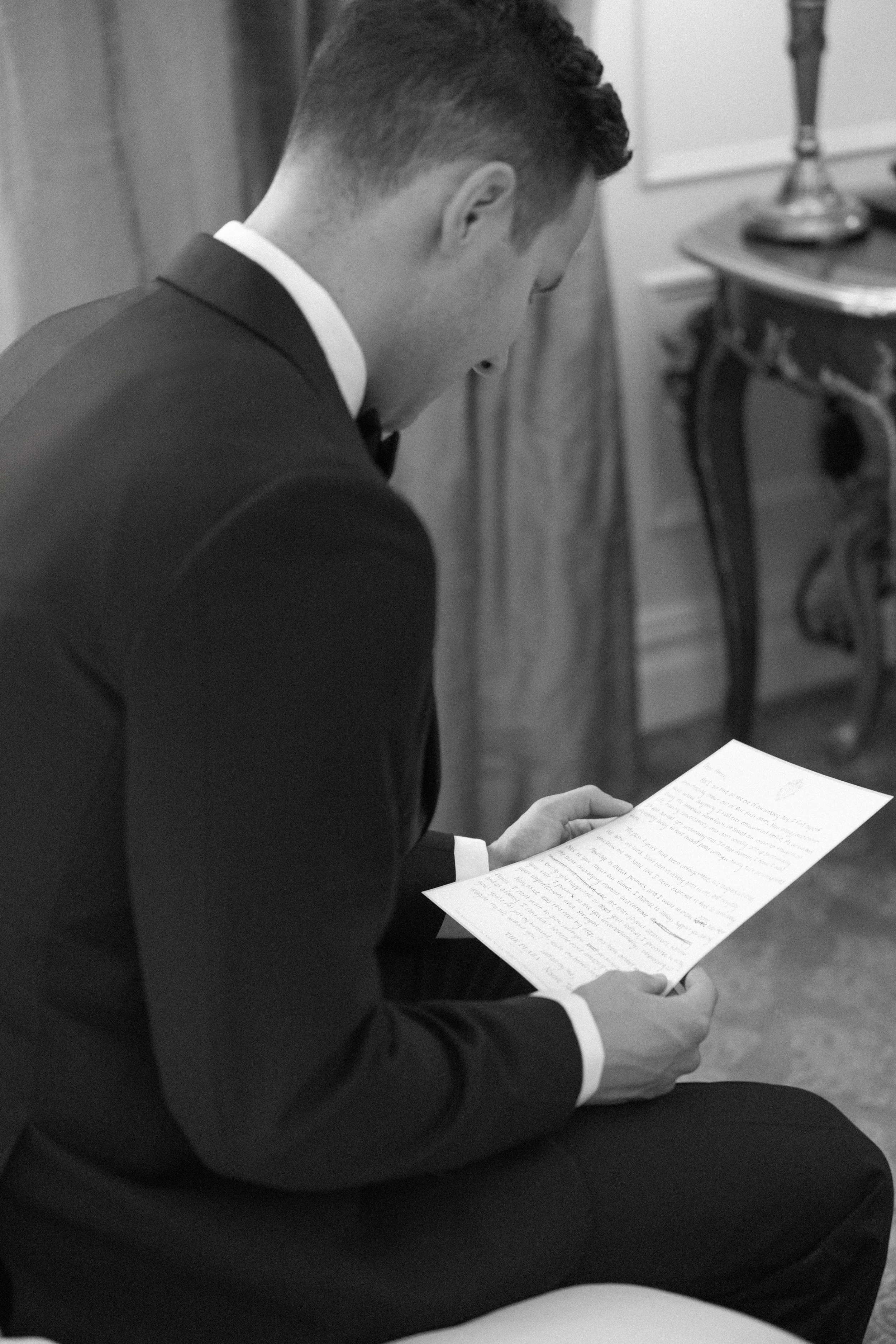 Man in formal attire reading a document in a room with classic furniture.