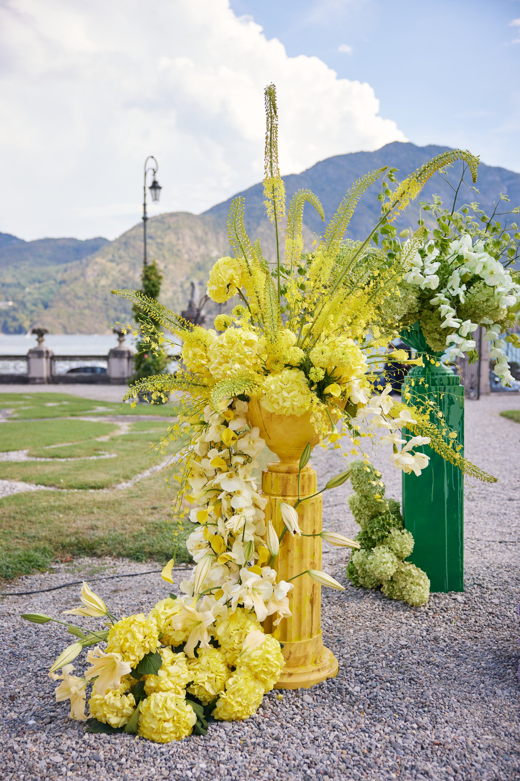 Yellow and white floral arrangement in a decorative vase outdoors with mountains and a cloudy sky in the background.