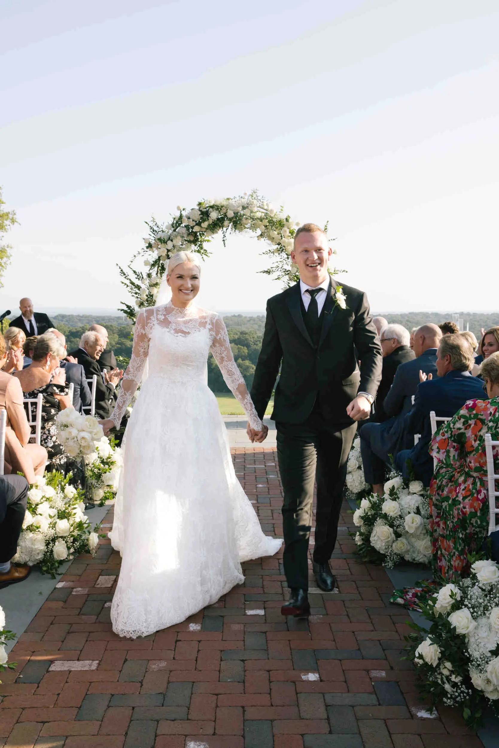 A bride and groom smiling and holding hands walking down the aisle during an outdoor wedding ceremony surrounded by friends and family. The bride wears a lace wedding gown and the groom is in a black tuxedo with a white tie, with floral decorations on and around a floral arch.