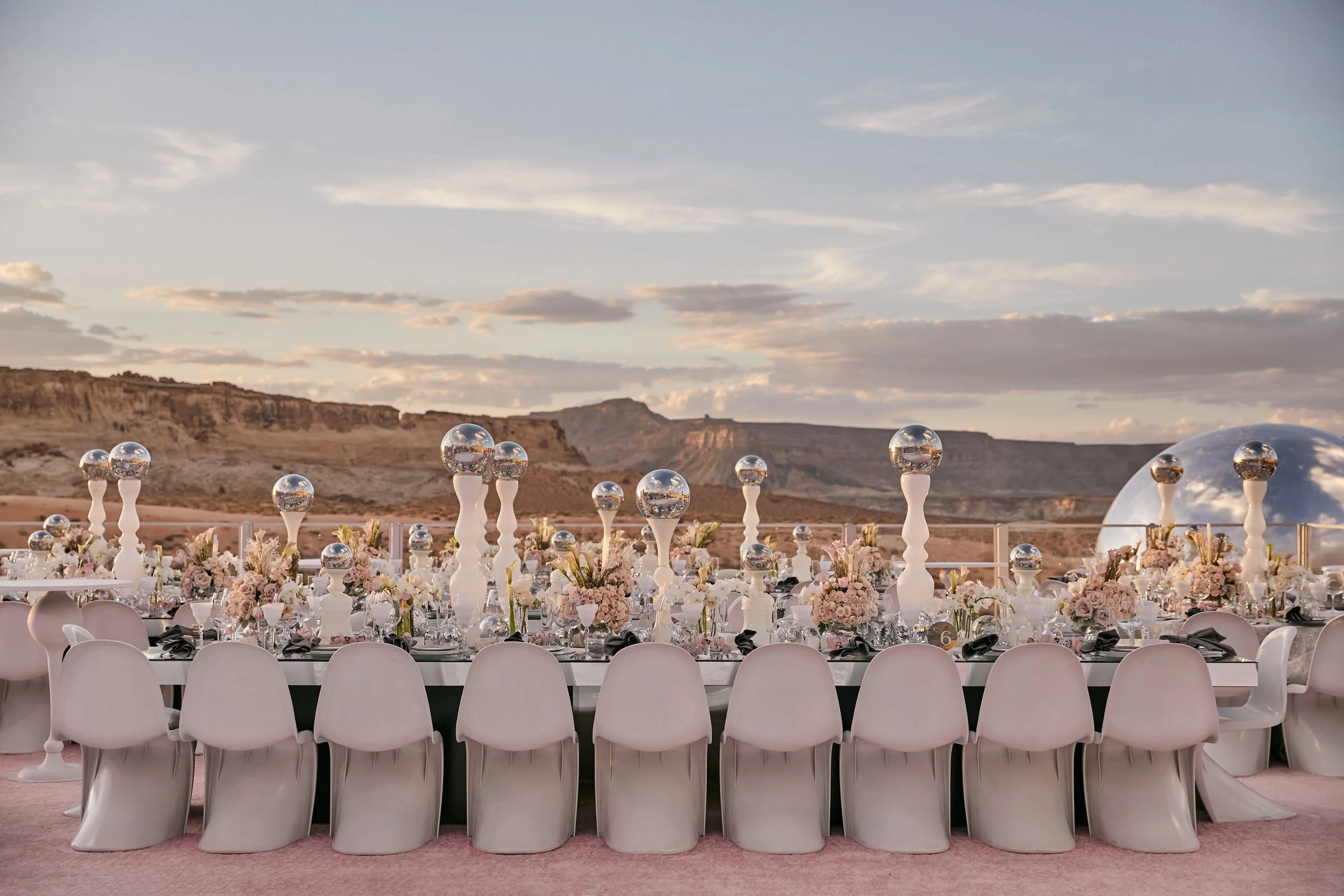 Elegant outdoor event setup with tables decorated with pink and white flowers and silver orbs on top, surrounded by white chairs, with a scenic landscape of cliffs and a cloudy sky in the background.