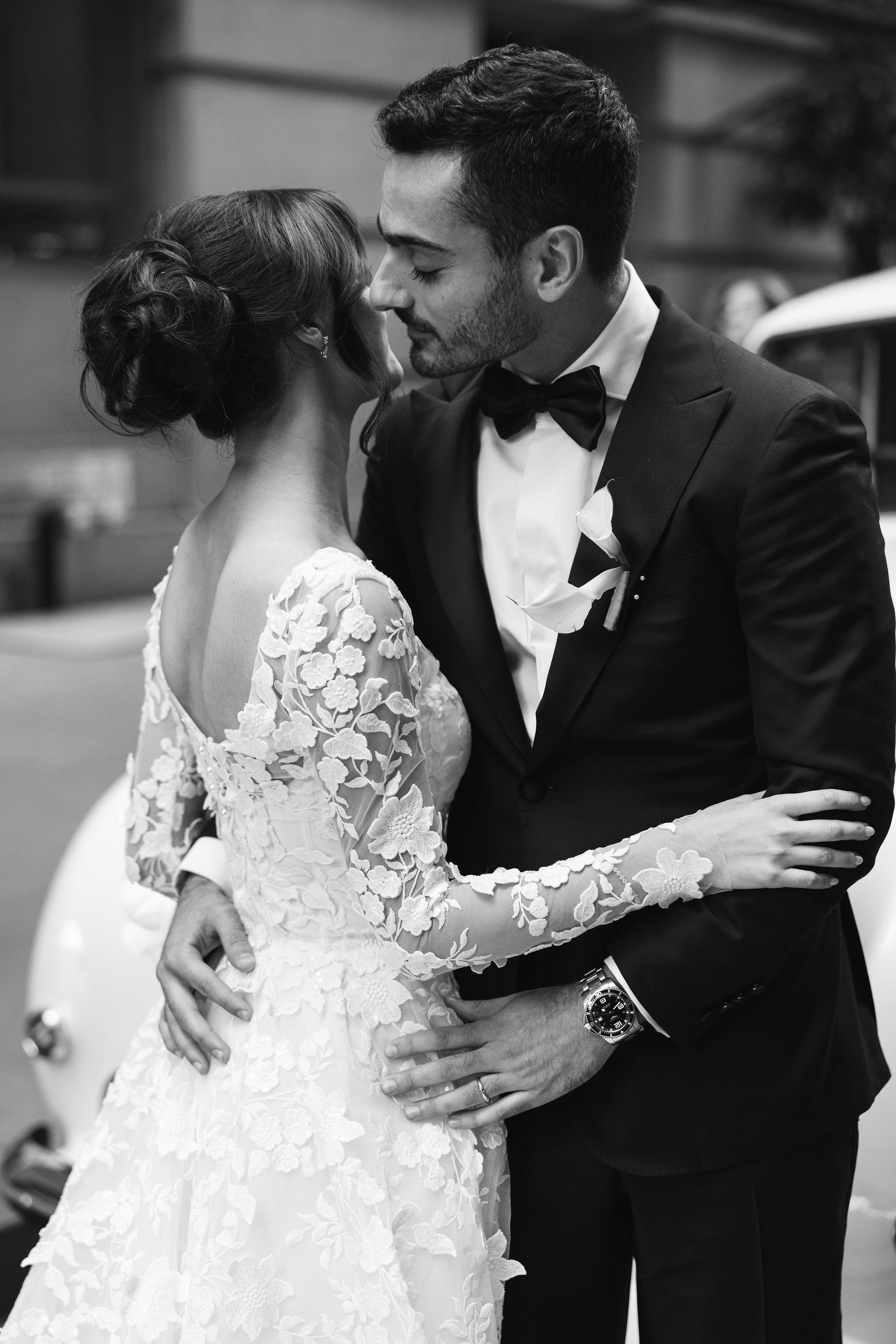 Black and white photograph of a newly married couple sharing a romantic moment, with the bride in a lace wedding gown and the groom in a tuxedo, embracing and touching foreheads.