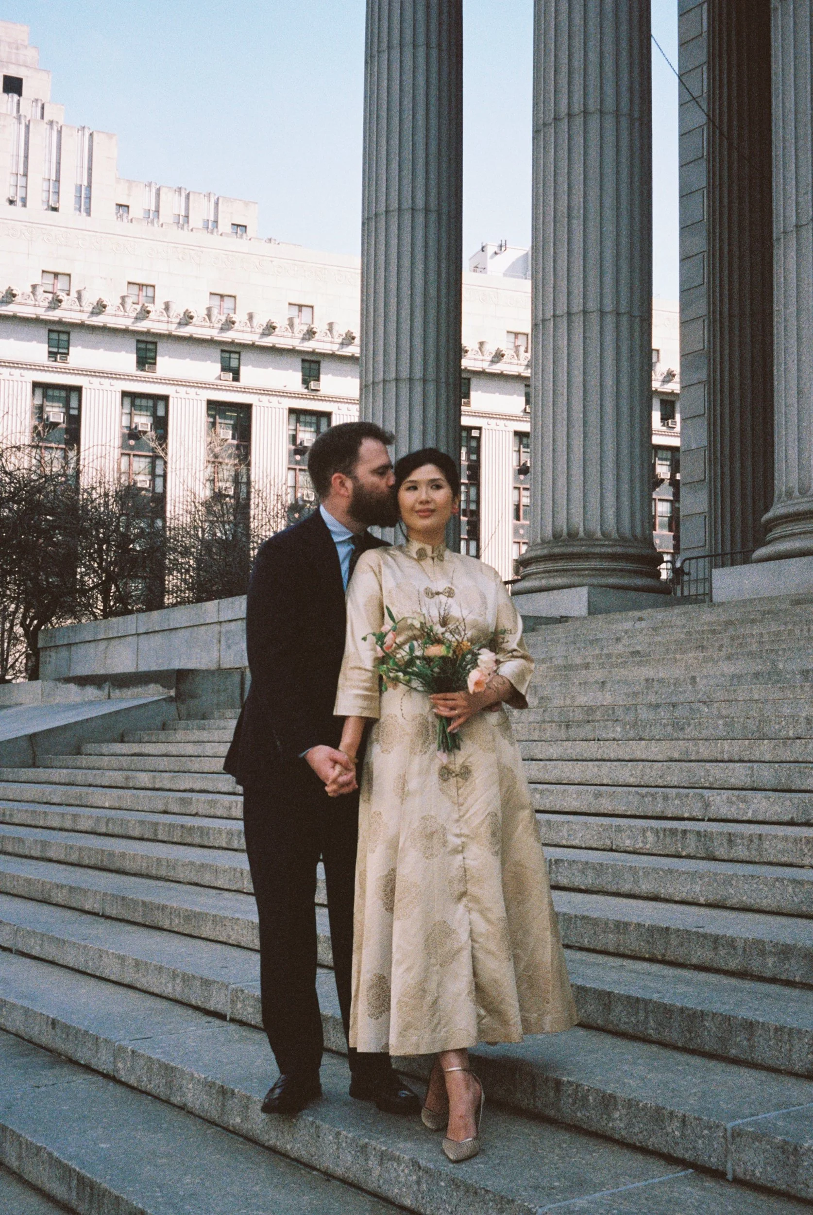 A man and woman standing on steps in front of a building with large columns, the man whispering to the woman, who is holding a bouquet.
