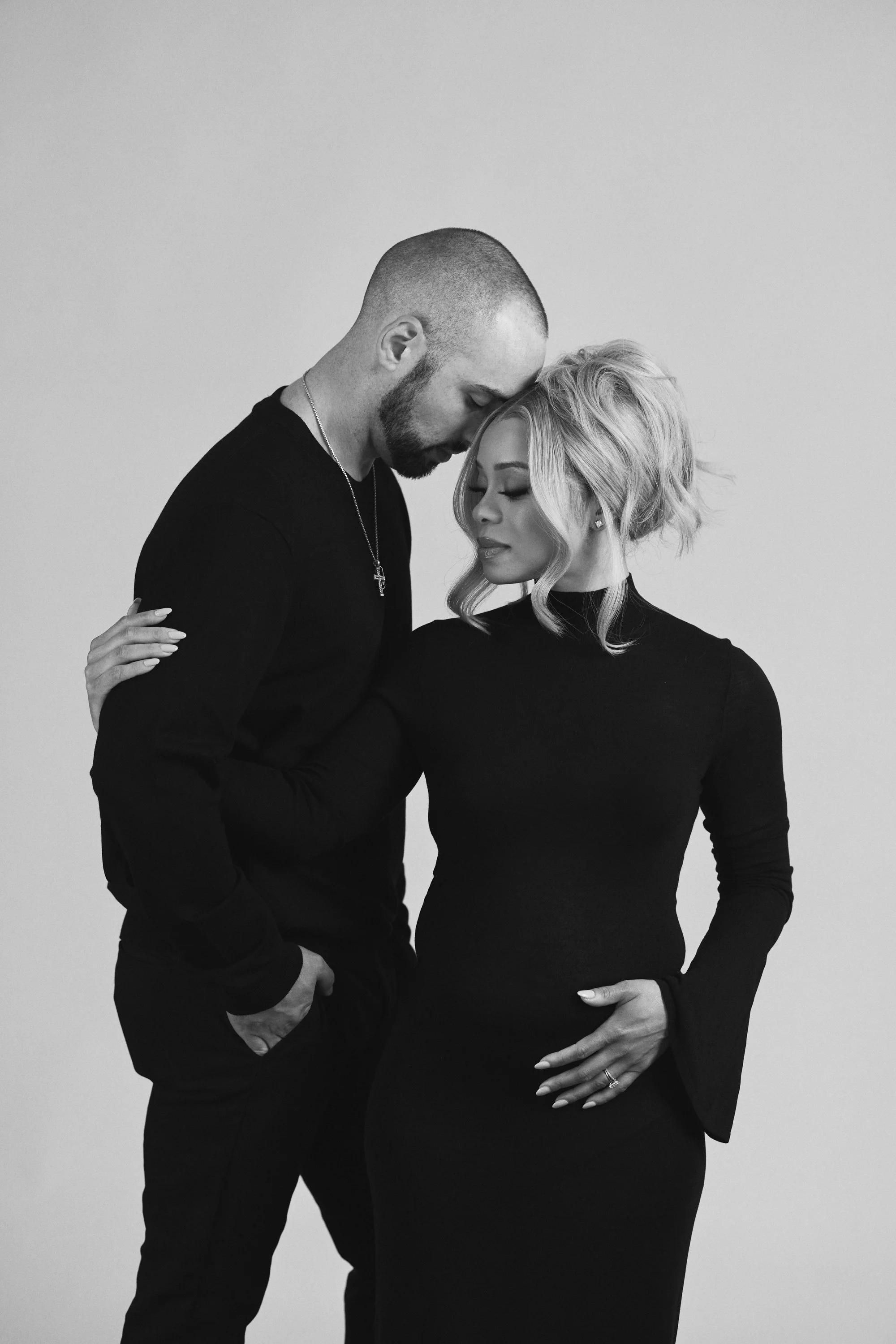 A black-and-white photo of a man and woman. The man has a shaved head, beard, and tattoos, and is dressed in black. The woman has wavy blonde hair, wears earrings, and a black long-sleeve dress. They are posed with their foreheads touching; the woman
