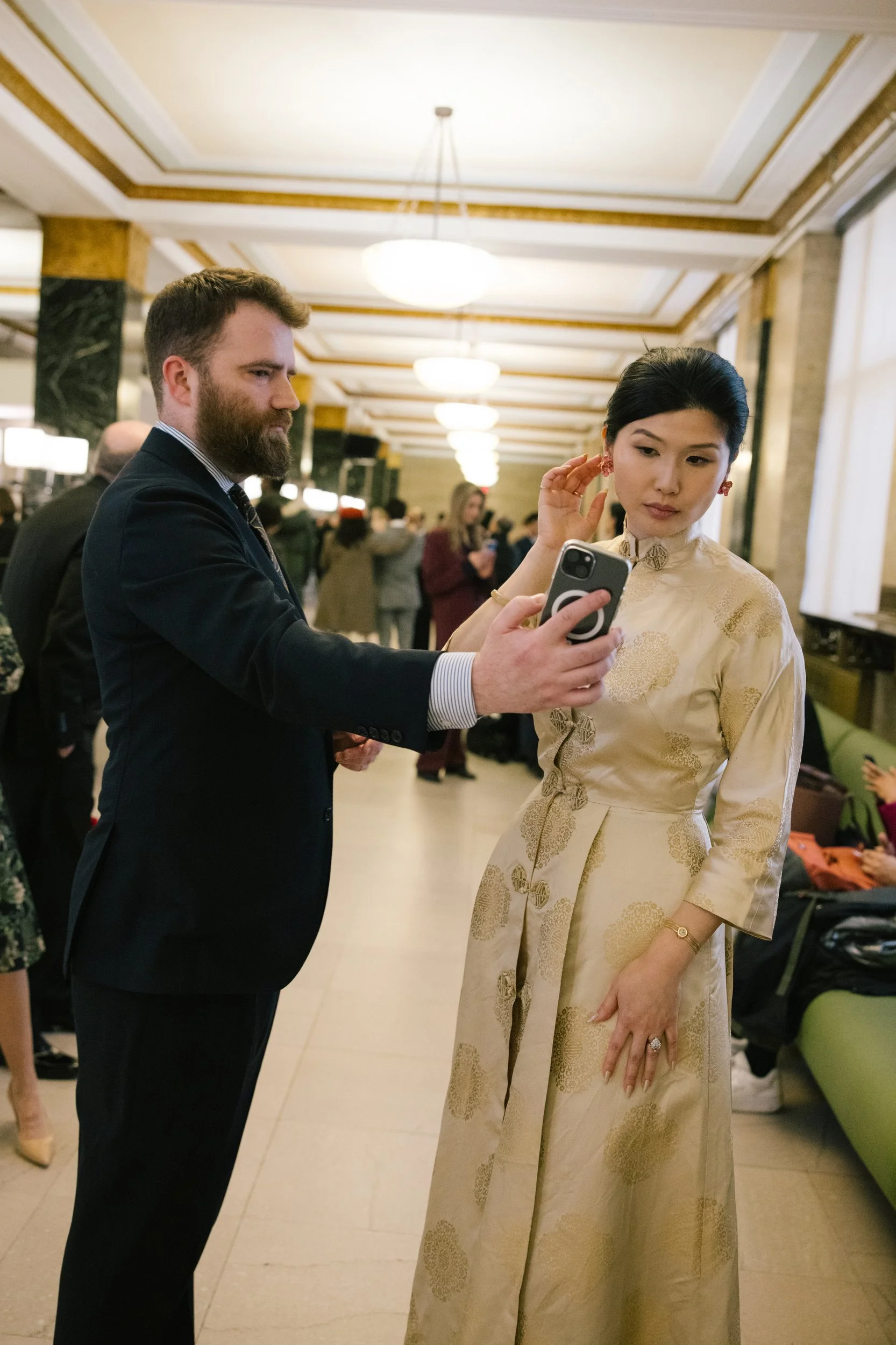 A man with a beard in a suit taking a selfie with a woman wearing traditional Vietnamese dress in a brightly lit, elegant indoor setting with many people in the background.