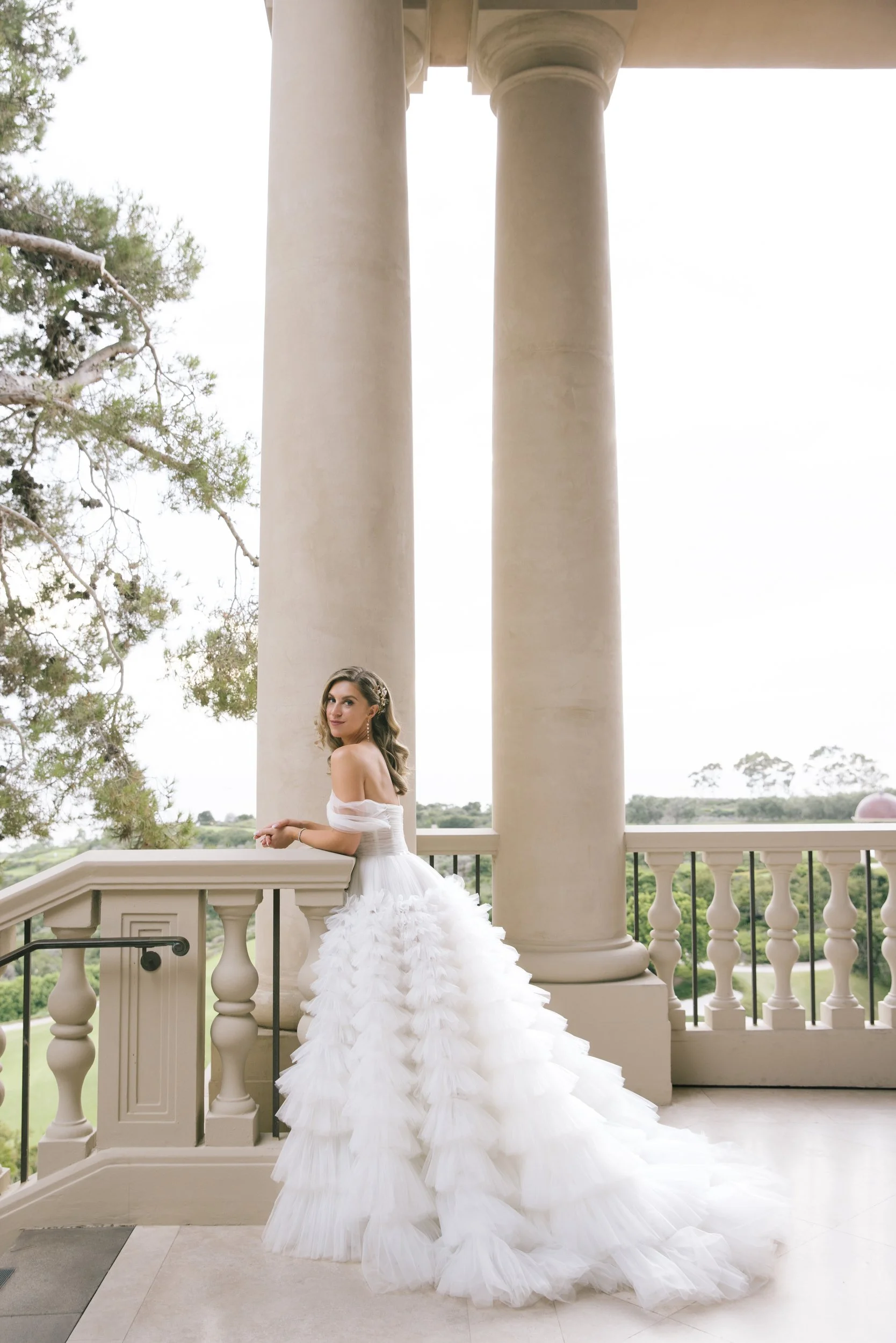 A woman in a white bridal gown with ruched bodice and large layered tulle skirt standing on a balcony with columns, overlooking a green landscape.