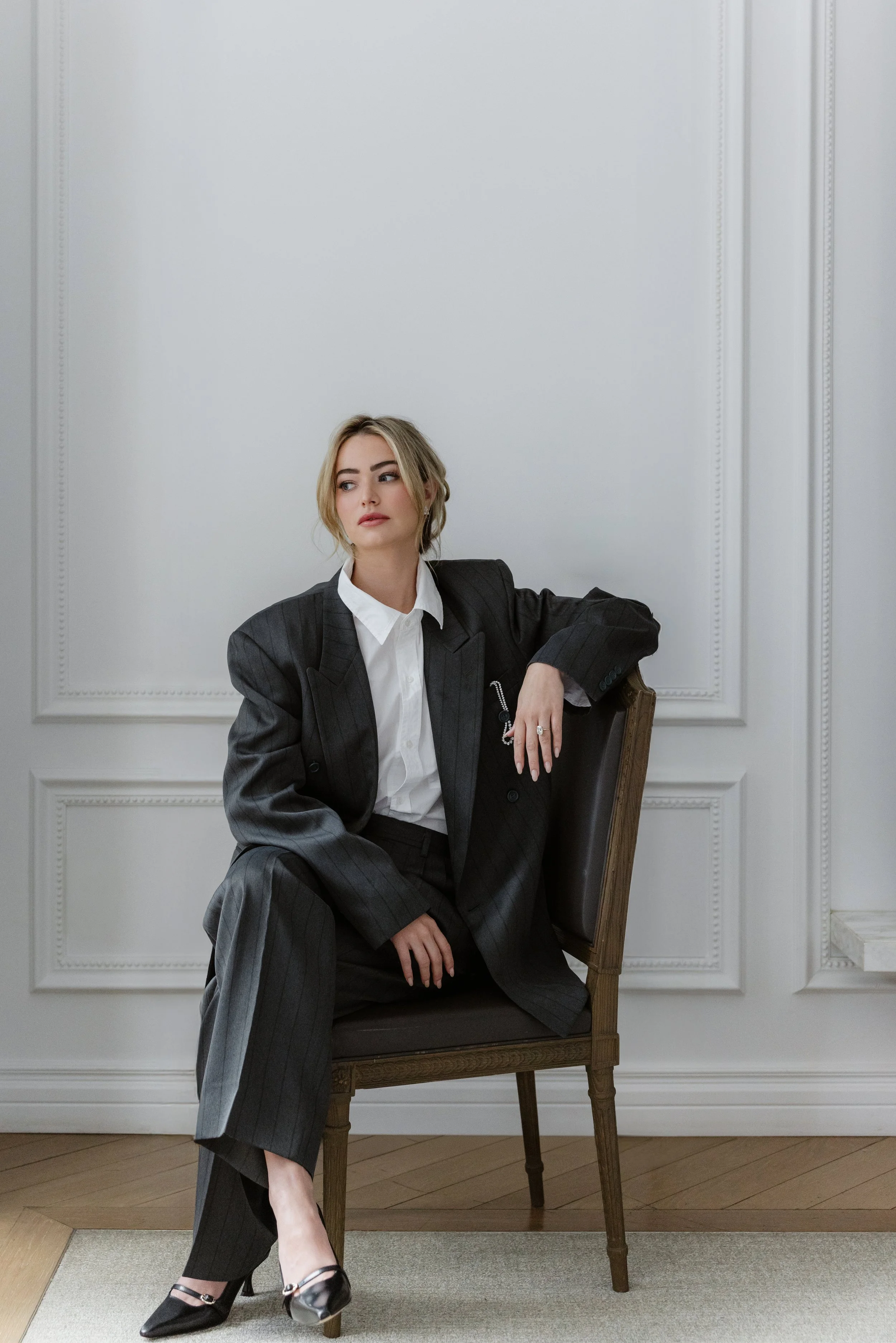 A woman in a gray pinstripe suit with a white shirt sits on a wooden chair, against a white wall with decorative molding. She has blonde hair and is looking to the side, with her left arm resting on the back of the chair. She wears black high heels w
