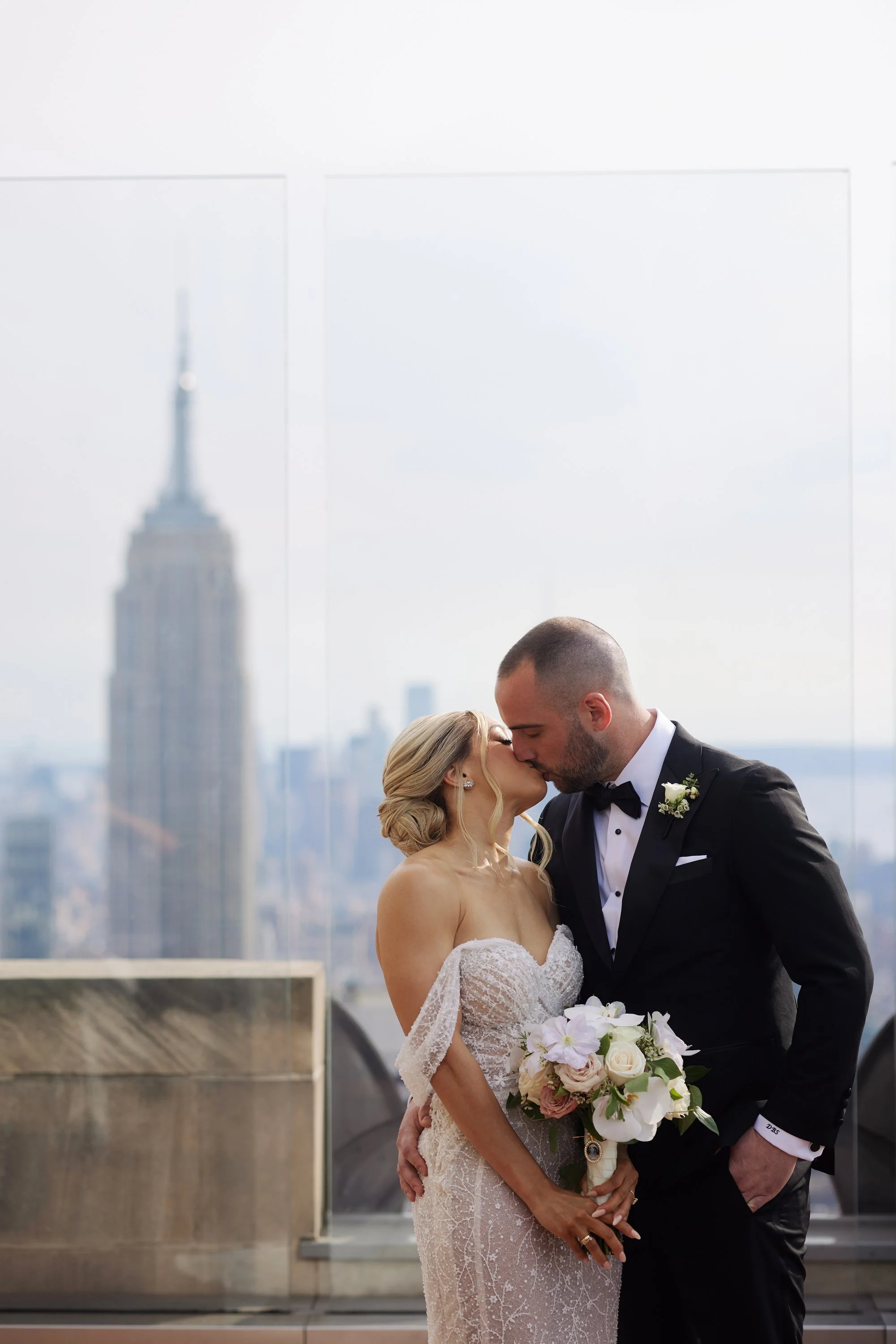 A bride and groom sharing a kiss on a rooftop with the Empire State Building in the background, holding a bouquet of flowers.