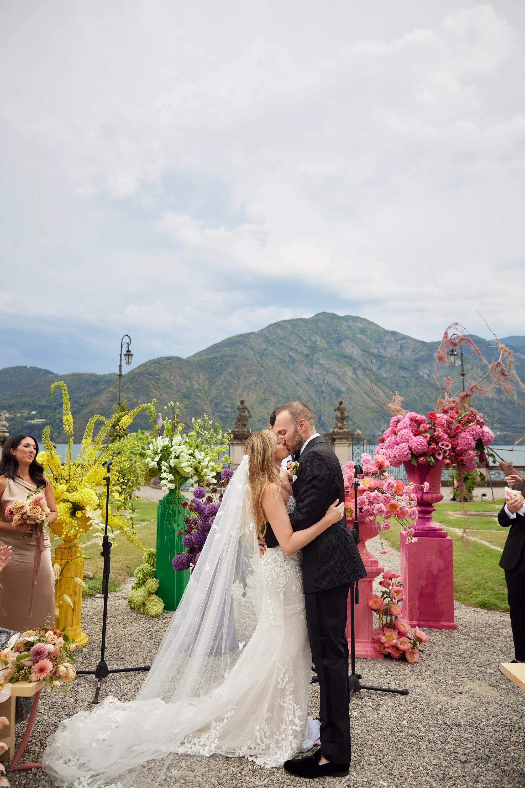 A wedding couple kissing during their outdoor ceremony, surrounded by colorful flower arrangements and mountains in the background.