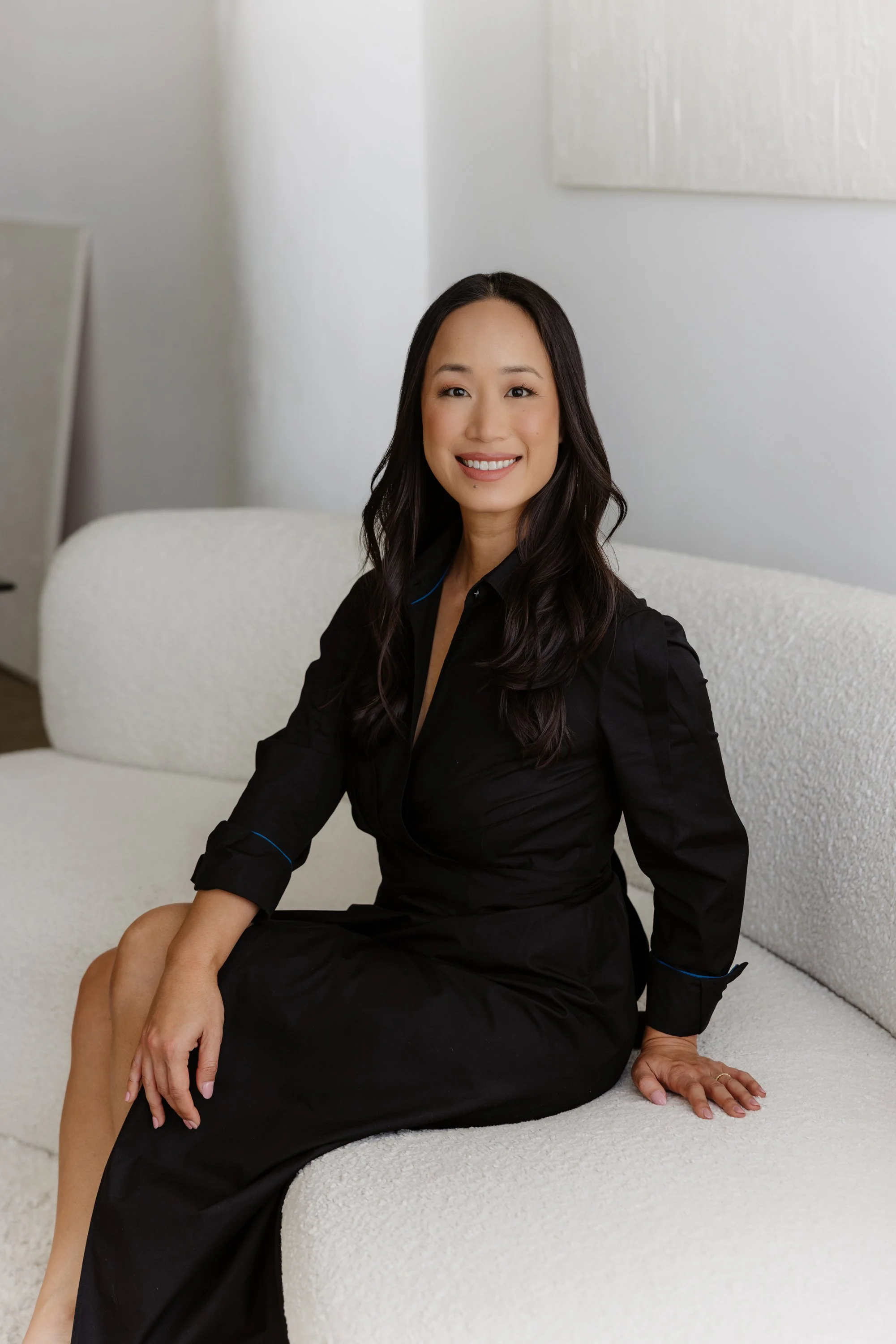A woman with long dark hair smiling while sitting on a white textured couch, dressed in a black dress, in a bright room with white walls and abstract art on the wall.