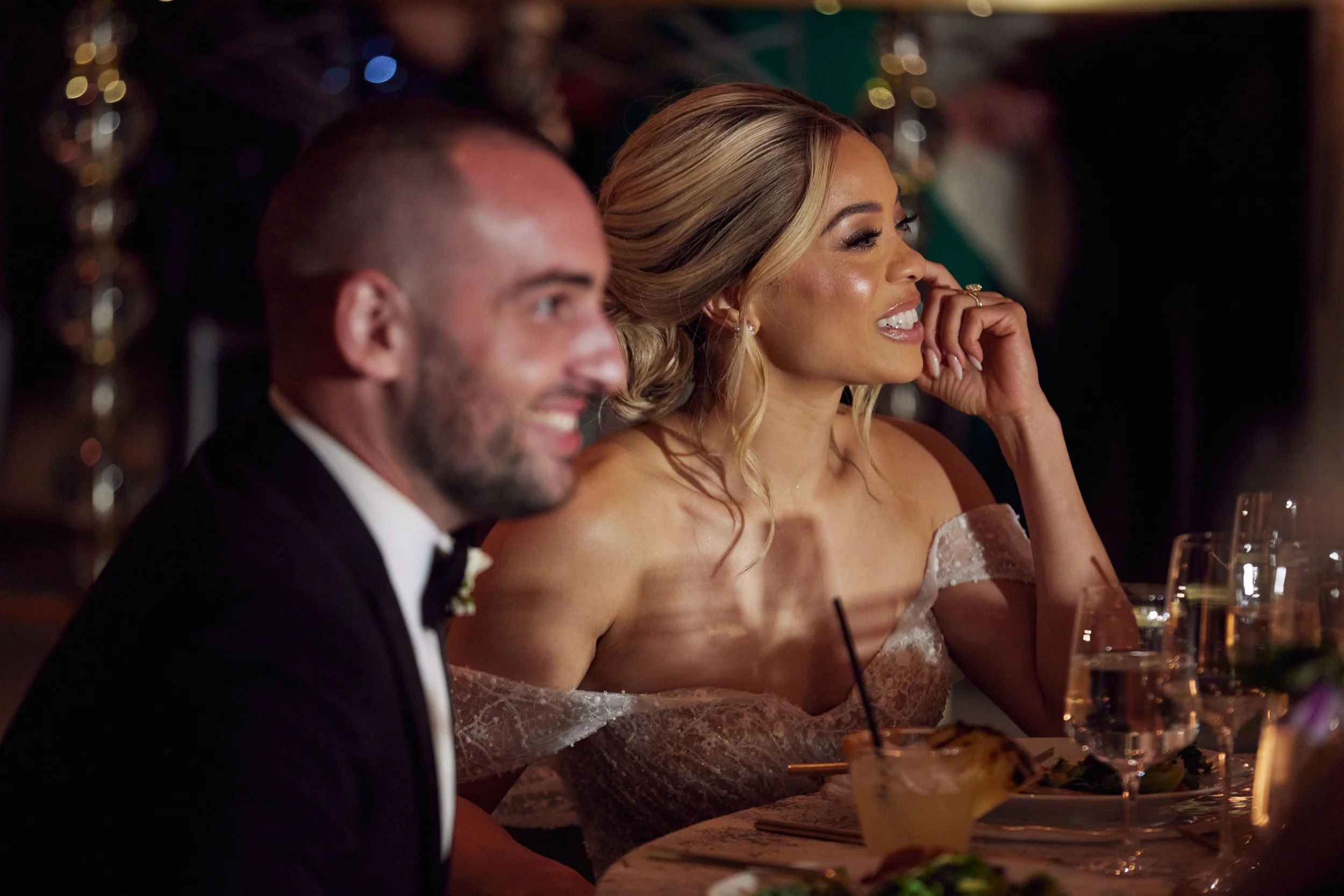 A joyful bride with blonde hair smiling and a groom with a beard and tuxedo dining at a wedding reception, with colorful bokeh lights in the background.
