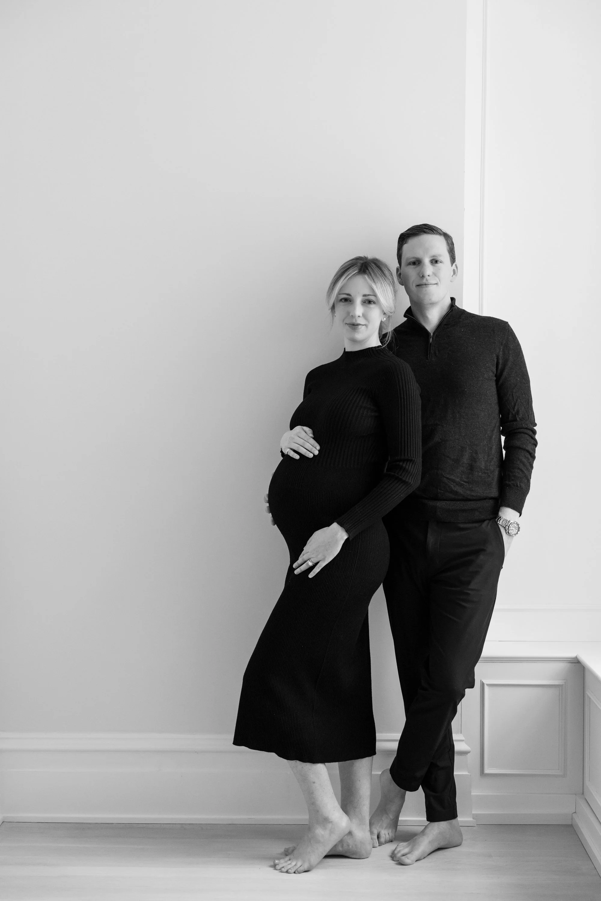 A pregnant woman and a man standing close together, both barefoot, with the woman holding her belly. They are dressed in dark clothing and standing against a plain wall, side by side.
