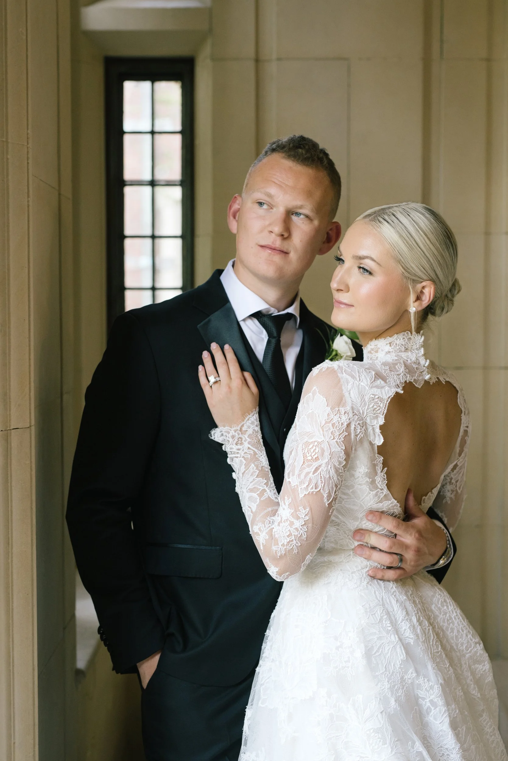 A bride and groom stand close together in a hallway, with the bride in a white lace wedding gown and the groom in a black tuxedo. The bride's hand rests on the groom's chest, showing her wedding ring, and they look at each other with gentle expressions.