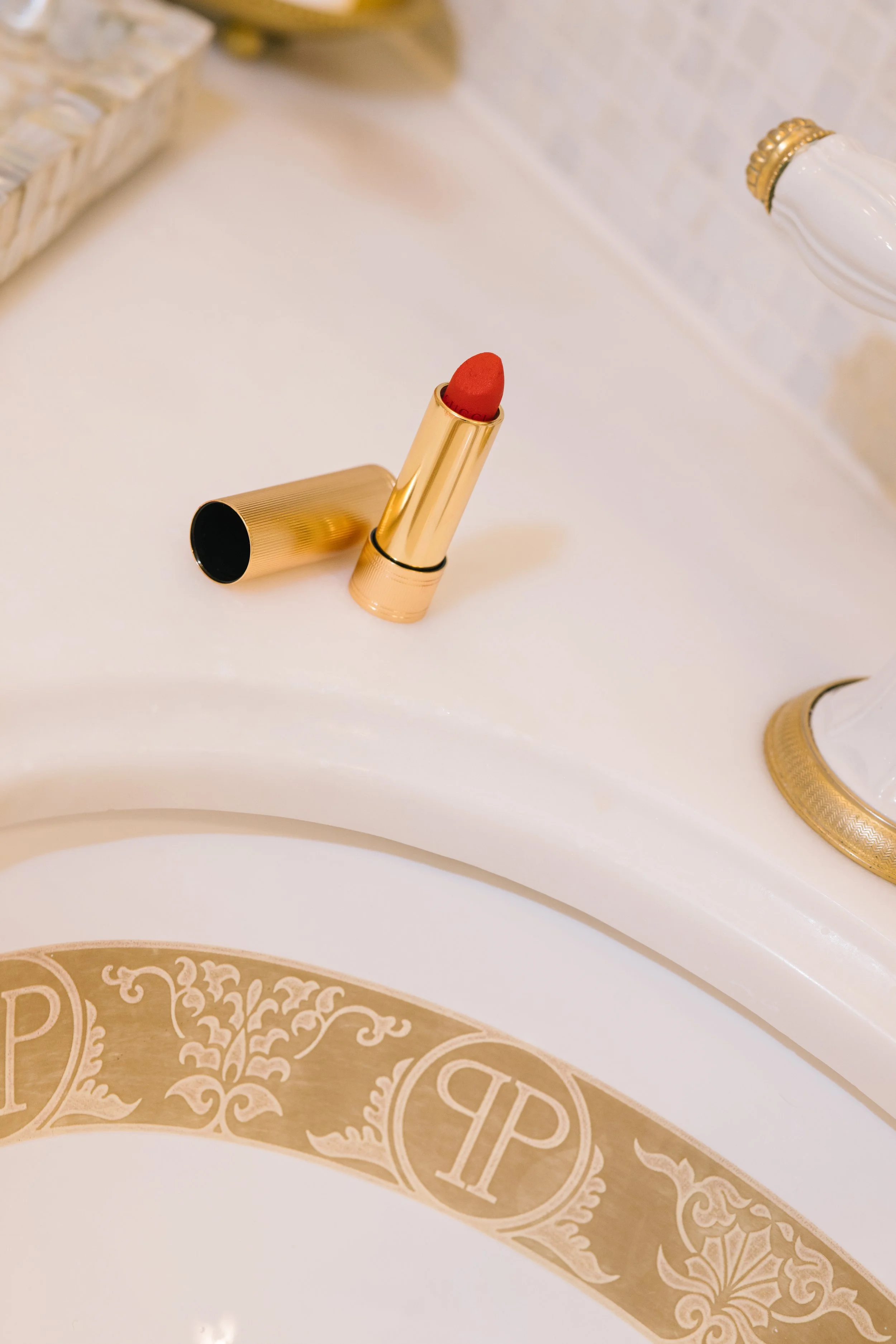 Open gold lipstick tube with red lipstick on a white surface, with ornate decorative paper underneath.
